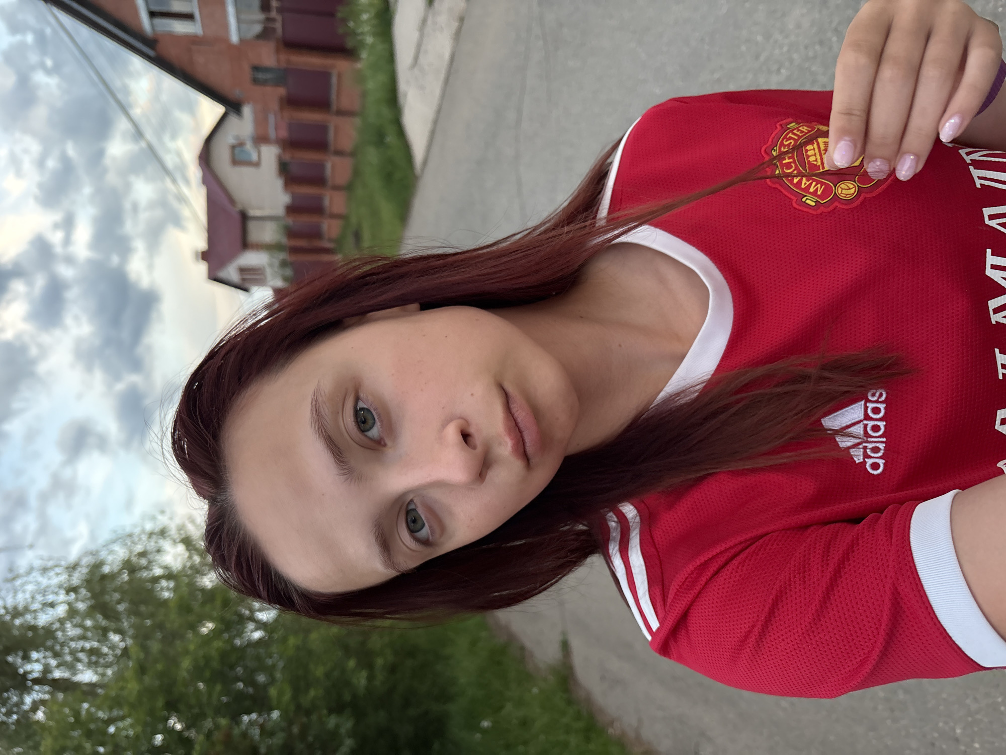 A young woman with fair skin and auburn hair is the central subject of the image. She's wearing a red Adidas jersey with white trim, featuring the Manchester United logo on the chest. The setting appears to be a residential area, possibly in Прикамский, Russia, as indicated by the presence of brick houses and lush greenery in the background. The woman is standing on what seems to be a paved road or path. The weather appears overcast, judging by the cloudy sky.