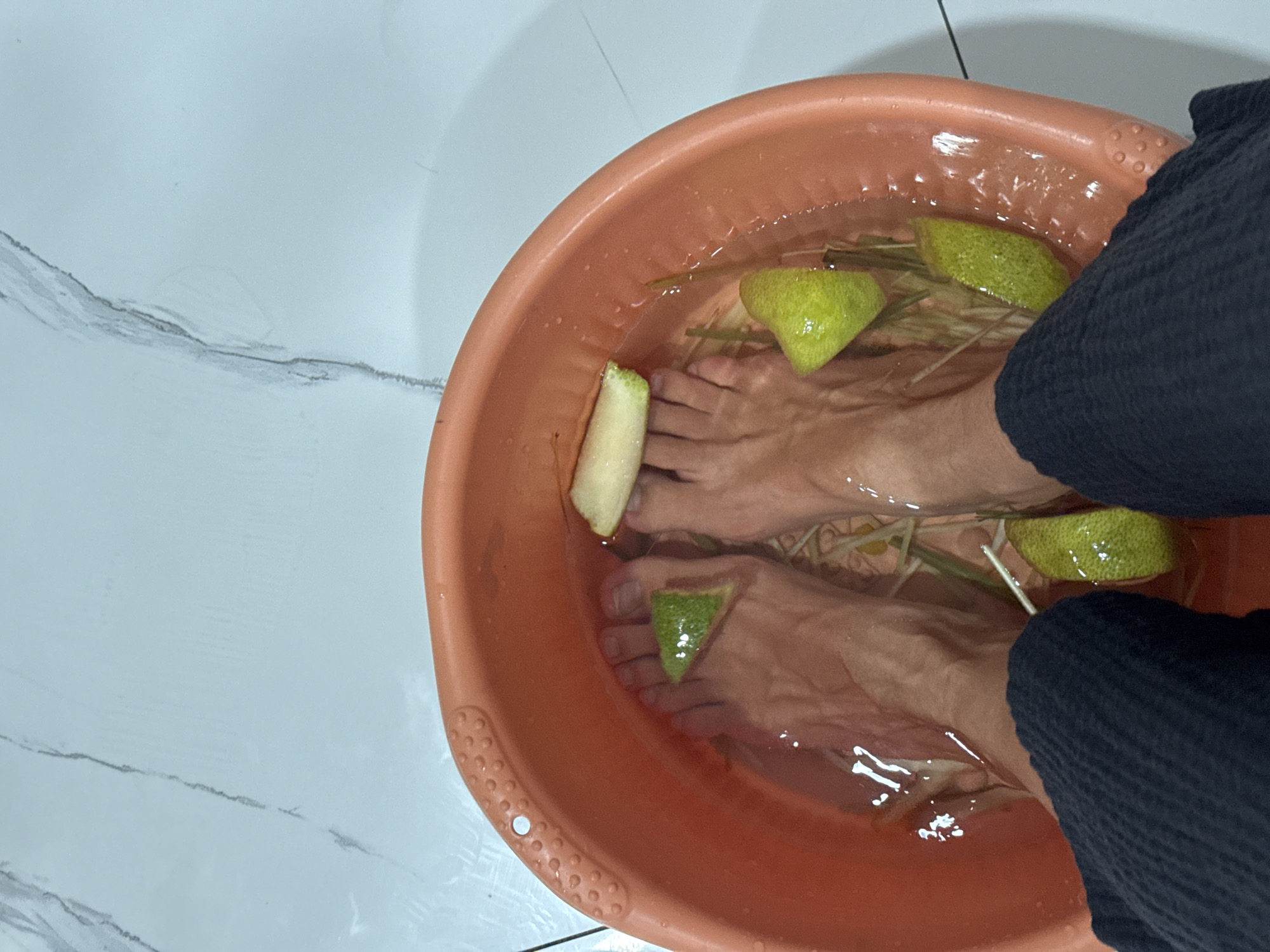 The image shows a high-angle shot of a person soaking their feet in an orange plastic tub filled with water, green citrus fruit slices, and lemongrass stalks. The feet are submerged in the water, with slices of citrus placed around them. The person is wearing dark blue trousers. The tub is placed on a white tiled floor with gray streaks. The location is indoors, but the city and exact place are unknown.