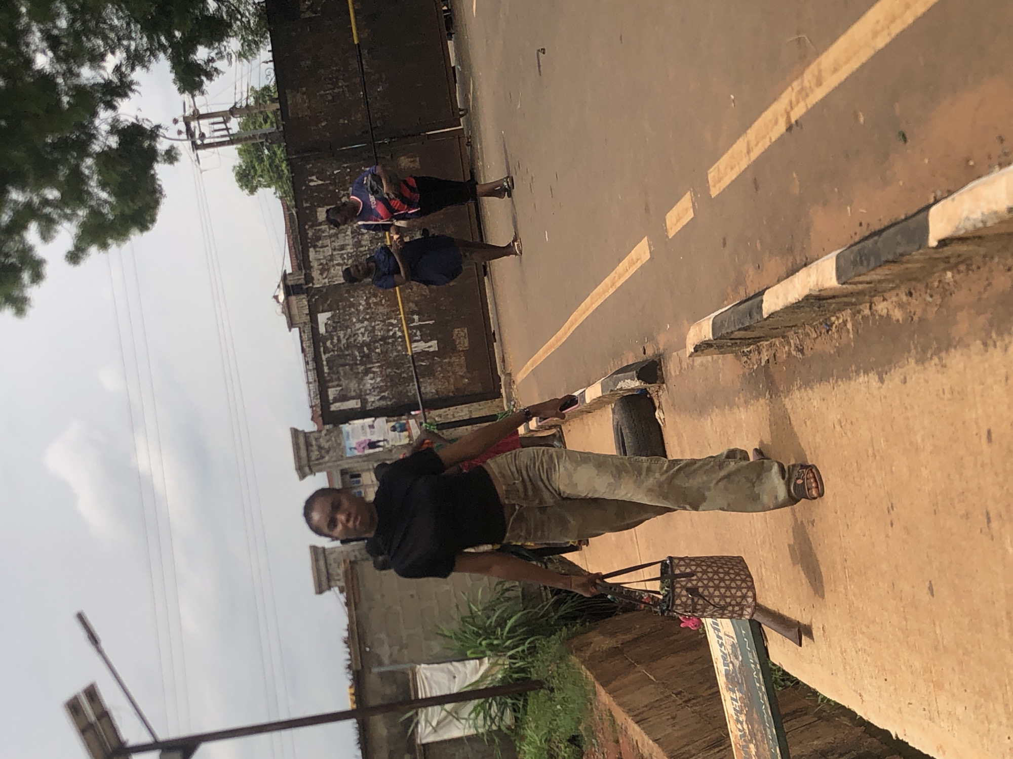 The image, rotated 90 degrees clockwise, captures an outdoor street scene in what appears to be a community in Nsugbe, Nigeria.
In the foreground, a young man stands on an earthen/dirt shoulder next to a paved road. He wears a black short-sleeved t-shirt, camouflage-patterned trousers, and sandals. He holds a distinctive woven bag with a brown and beige geometric pattern in his right hand, while his left hand holds a phone. He looks directly at the camera with a neutral expression.
To his right (if correctly oriented), a paved road stretches into the distance, marked with a faded yellow dashed line. A black and white painted curb separates the road from the shoulder. Further along the road, two other individuals are visible. One person wears a blue and red striped top with dark bottoms, carrying a bag. The other wears a dark blue dress or top with dark bottoms. They are standing by a weathered, possibly concrete or brick, wall that runs parallel to the road.
The background features a bright, partly cloudy sky. Lush green tree foliage is visible on the left side of the frame. Utility poles and overhead power lines crisscross the sky. A solar-powered street light fixture is present in the lower left section. On the embankment near the foreground subject, there are some green bushes and a discarded tire.
Partially visible text on a sign or structure in the bottom right of the image reads "...ISTRIBUTION DEPARTMENT". The overall lighting suggests it is daytime, likely during clear to partly cloudy conditions.