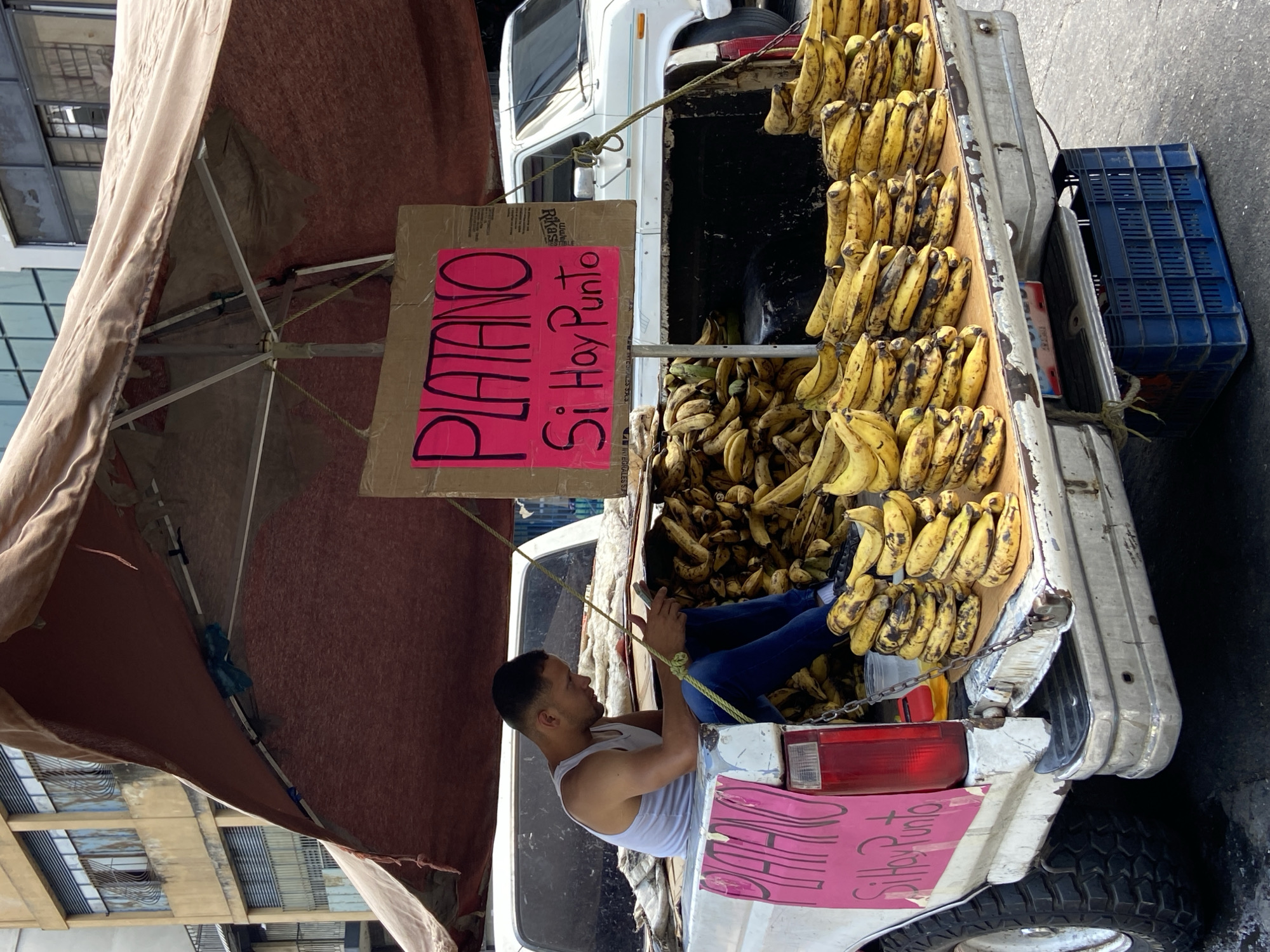 The image displays an urban street vending operation in Caracas, Venezuela. A light-skinned adult male with short dark hair, dressed in a white sleeveless tank top and blue jeans, is seated on the open tailgate of a white, weathered pickup truck. He is focused on a small electronic device held in both hands. The truck bed is filled with a substantial quantity of ripe and ripening plantains, arranged in multiple stacks across the tailgate and piled higher towards the front of the bed.
A large, worn, reddish-brown fabric canopy, supported by a central metal pole and frame, extends over the truck bed, providing shade. A yellow rope, part of the canopy's tethering system, is visible near the seated individual. Attached to the canopy structure is a prominent cardboard sign, handwritten with "PLATANO" in large pink capital letters and "Si Hay Punto" in black script below, indicating that plantains are for sale and card payments are accepted. A smaller, identical sign is affixed to the exterior of the truck's tailgate, partially obscured. Beneath the open tailgate, a blue plastic crate rests on the ground.
In the background, a multi-story building with multiple windows and metal security bars is visible, showing signs of age and weathering. Part of another white vehicle is visible on the right side of the frame, parked further down the street. The ground is dark, appearing to be asphalt or concrete with some visible moisture.