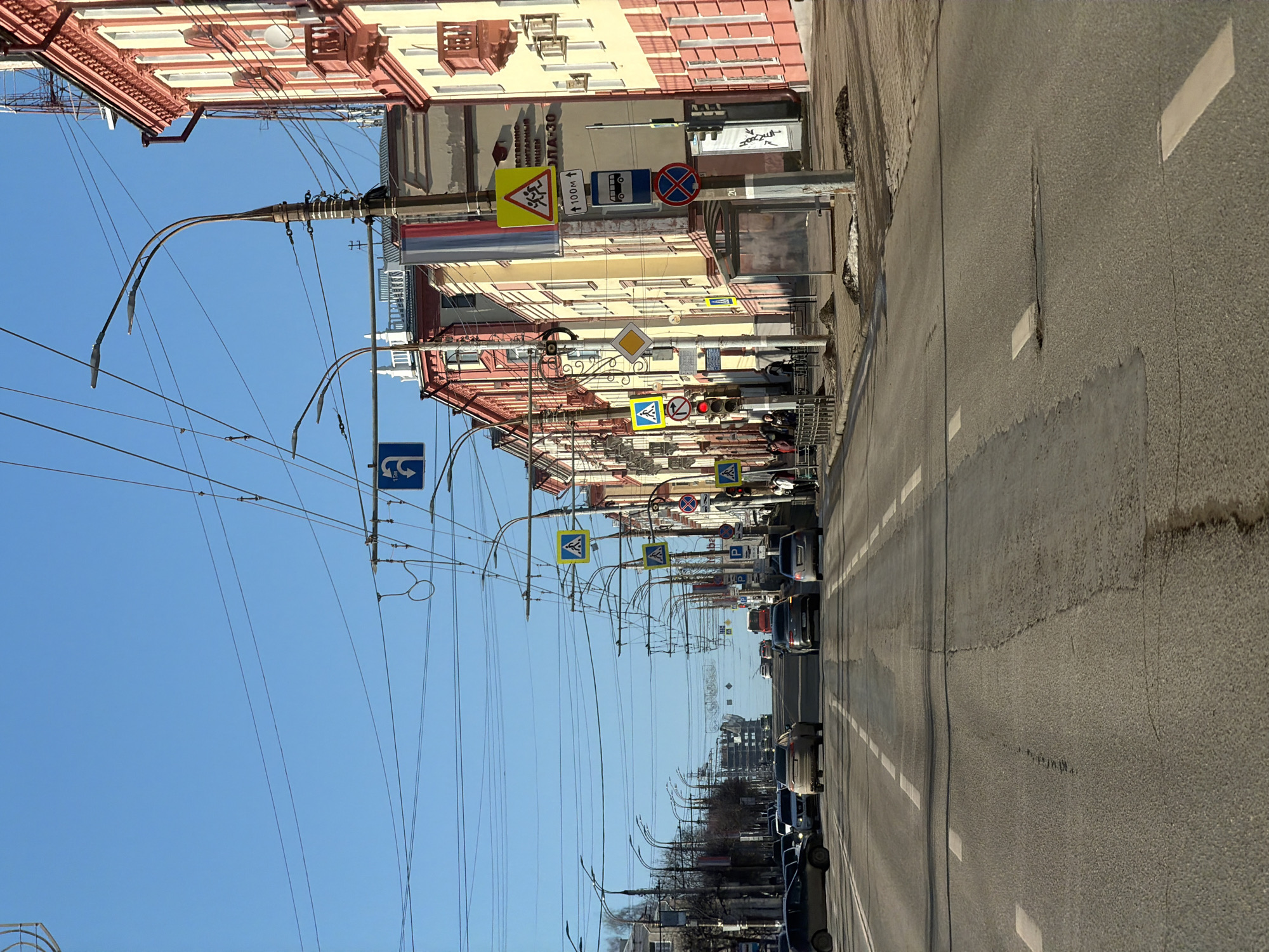 The image shows a street in Izhevsk, Russia. On the right, cars are parked along the road. Utility poles line the left, with a network of wires stretching overhead. Street signs are visible along the buildings. The buildings are a mix of light yellow and red brick. The sky is clear and blue.