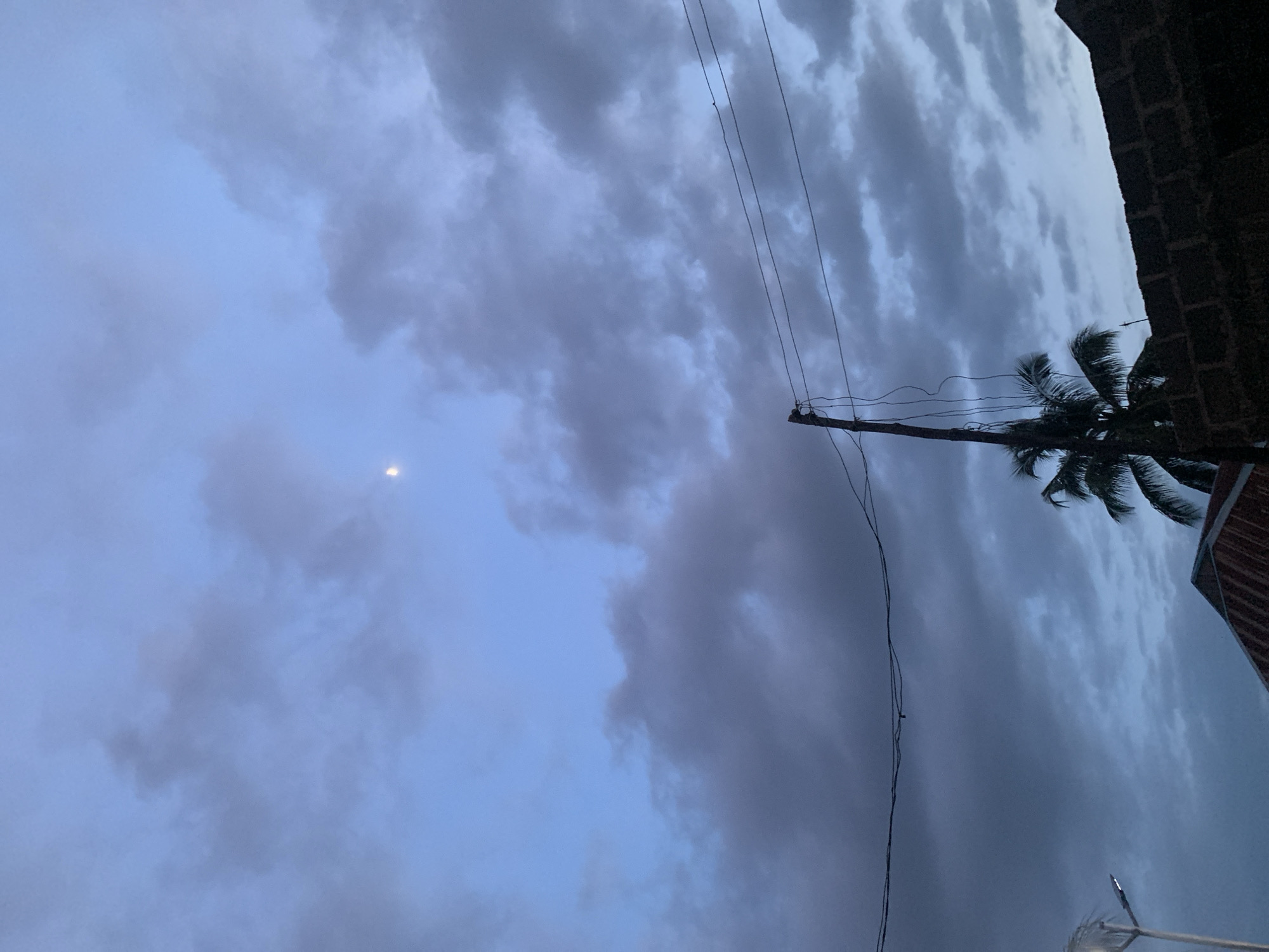 This is a low-angle, medium shot of the sky over Sabon Tasha, Nigeria, taken during twilight. The sky is a deep blue with scattered dark gray clouds. A sliver of the moon is visible in the upper left quadrant of the image. In the foreground, a telephone pole stands to the left of center, with several wires running horizontally across the frame. To the right of the pole, a dark brick wall is visible, and behind it, the peak of a building with a reddish-brown roof is peeking out. To the left of the pole, a street lamp with a white light is visible, and to the right of the pole and peeking over the brick wall is the frond of a palm tree. The overall scene suggests a dusk or pre-dawn setting.