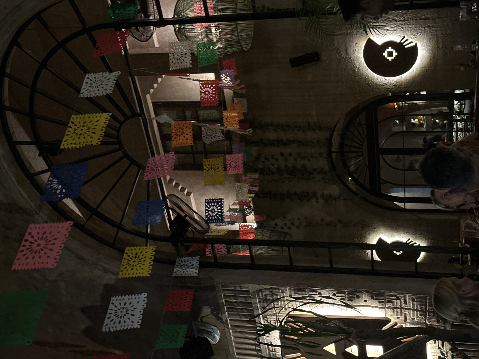 The image is a low-angle shot taken at night in what appears to be a restaurant or bar in Canggu, Indonesia. The main visual element is an iron-wrought, arched structure adorned with colorful, decorative paper flags or banners, typical of Mexican “papel picado” art. A staircase rises in the background, partially obscured by the decorations. A black fan is affixed to the iron structure.
To the right of the image, round windows with ornate frames are visible, and through one of them, a pool of water can be seen. Two decorative lights are mounted on the wall; one is round with a central design, and the other is shaped like a bat with glowing eyes.
In the bottom-right corner, part of a table is visible with a few people seated around it. Their faces are partially obscured by shadows, but they appear to be engaged in conversation. Plants are also visible. The lighting is dim, and the overall atmosphere suggests a casual, nighttime establishment. The image suggests a bohemian aesthetic typical of some venues in Canggu, known for its international dining and nightlife scene.