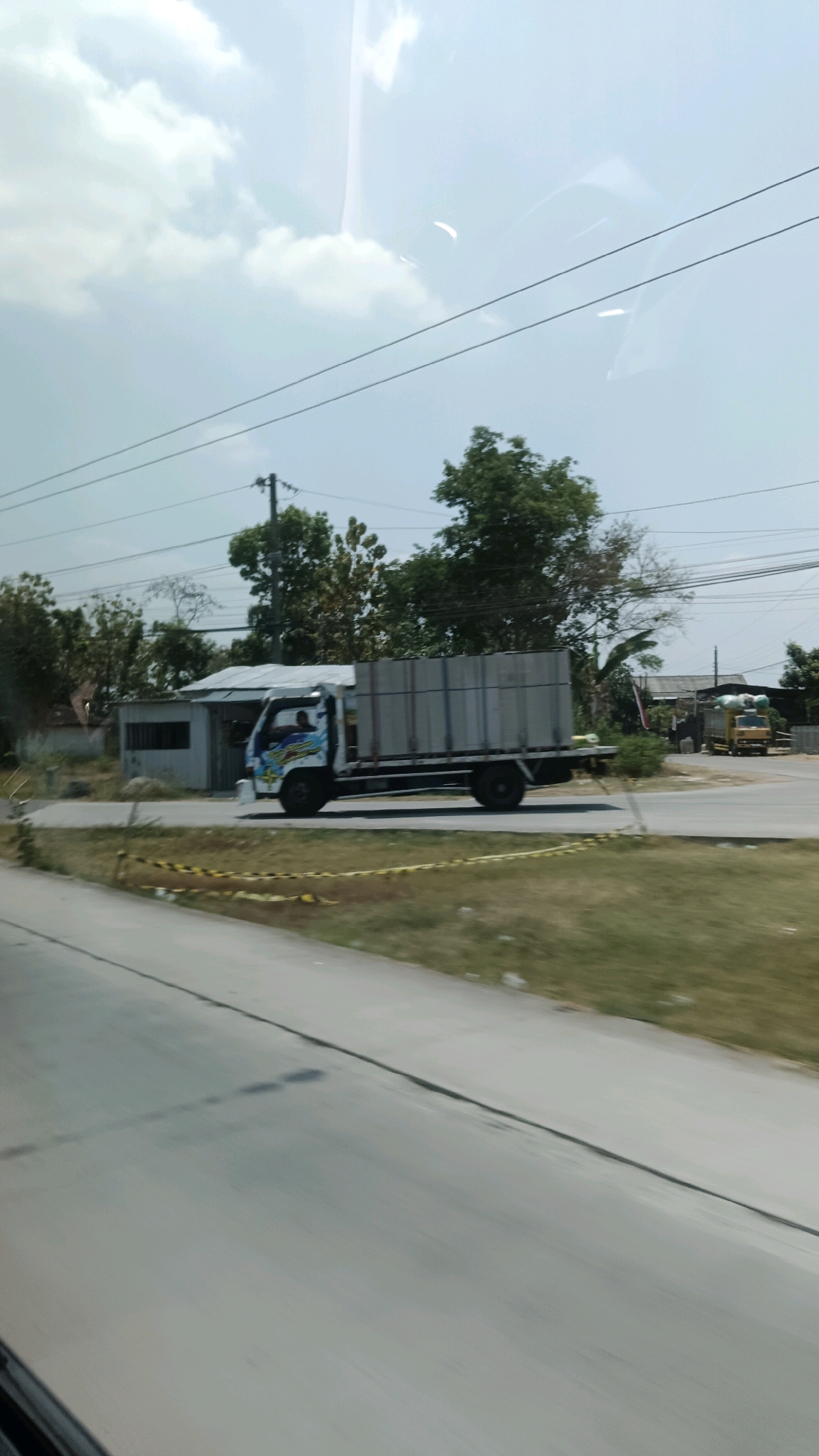 The image depicts a street scene in Ngarum, Indonesia, on a cloudy day. The foreground shows a paved road with a grassy shoulder bordered by yellow and black caution tape. In the midground, a white and blue truck with a large, gray cargo container is driving on the road. The truck has a driver visible in the cabin. Behind the truck, there are several buildings with corrugated metal roofs, some obscured by trees. In the background, more trees and power lines are visible, along with another yellow truck in the distance. The overall environment suggests a rural or semi-urban setting.