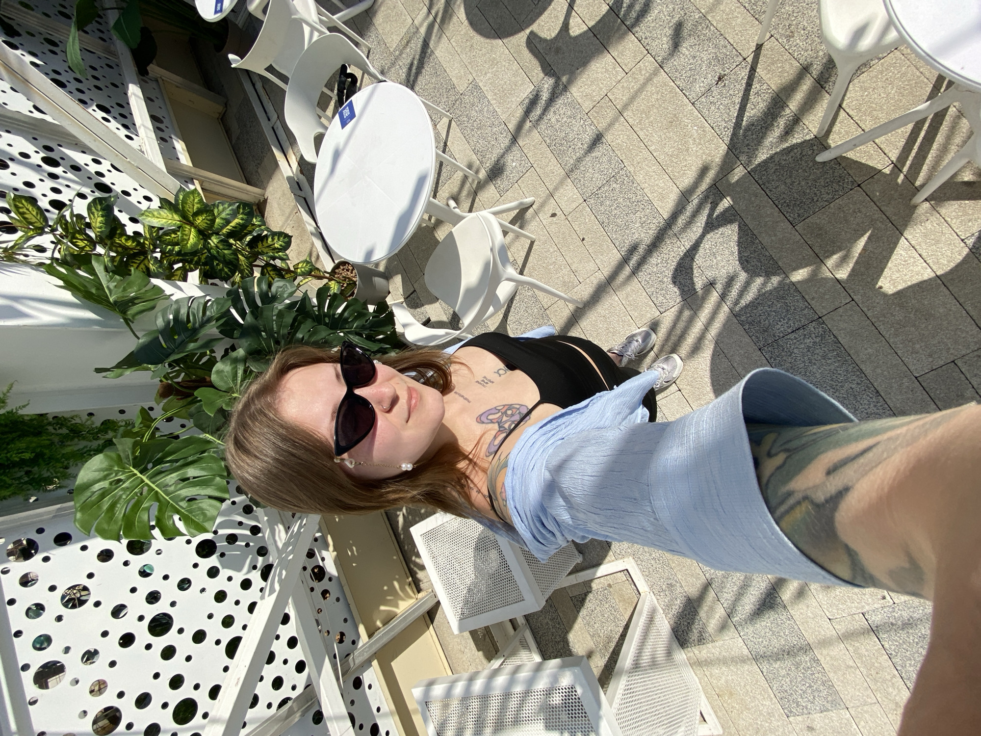A woman with sunglasses and tattoos is taking a selfie outdoors. She's wearing a black top and a light blue shirt. The location appears to be a patio or outdoor seating area with white tables and chairs. Green plants and decorative structures with circular cutouts are visible in the background. The ground is paved with grey brick. The image suggests the location could be in Saratov, Russia, although this is not definitively confirmed by visual cues.