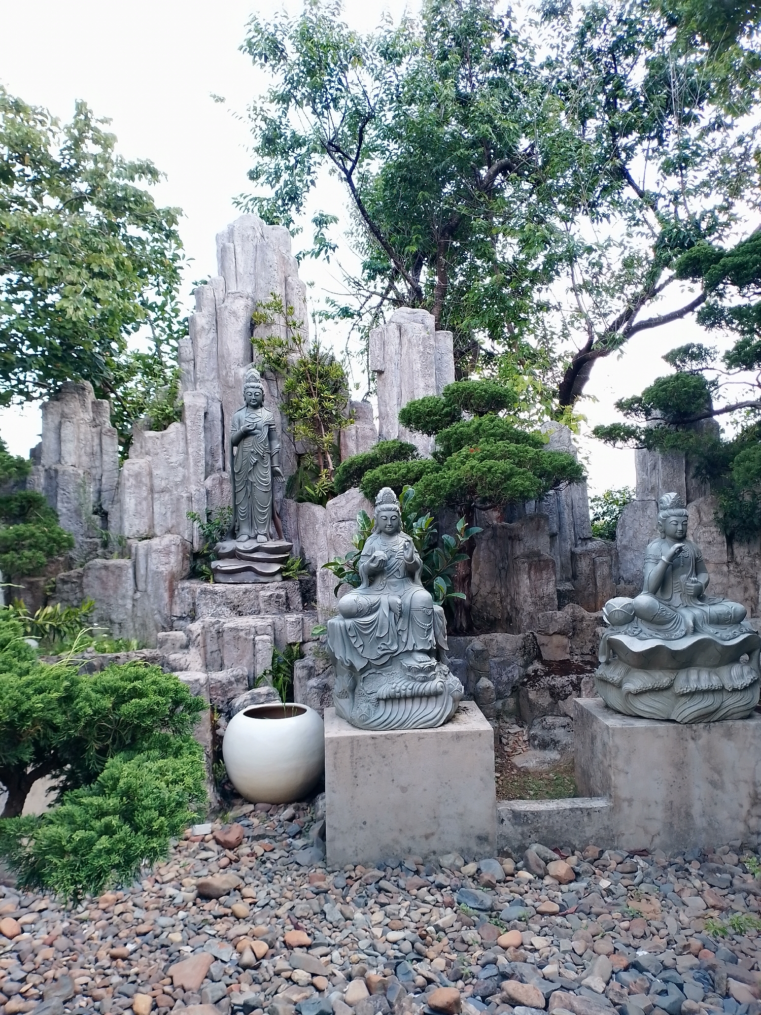 The image depicts an outdoor garden or park setting in Thôn 5, Vietnam, featuring three Buddha statues of differing designs positioned against a backdrop of artificial rock formations and foliage. The central statue, in a seated position, rests atop a concrete base, as does a second seated statue to the right. The third Buddha, in a standing pose, is elevated on a rocky outcrop.
The ground is covered in small, colorful pebbles. There is a white ceramic pot to the left of the central statue. Lush green bushes and trees surround the statues and rock formations, creating a serene and contemplative atmosphere. The sky is overcast, providing soft, diffused lighting.