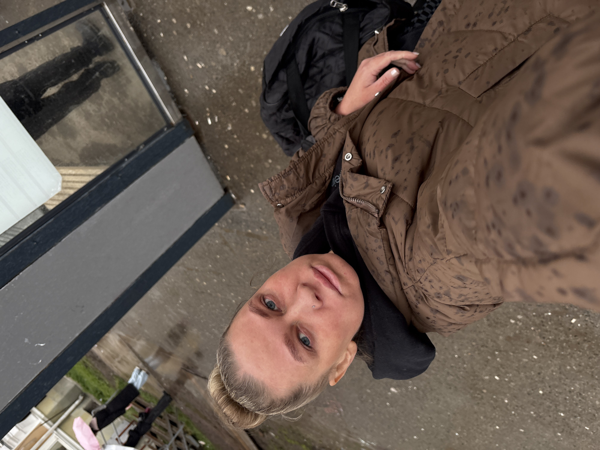 A woman with blonde hair pulled back into a bun and blue eyes, wearing a brown patterned puffer jacket and a black hoodie, is taking a selfie. She has a nose piercing. A black backpack is visible to her right. The photo appears to be taken outside, on a wet concrete surface. A glass window with a metal frame reflects a person's legs and shoes. In the background, there is a glimpse of green grass and white building structures. The reflective glass, the wet concrete, and the architectural details suggest an urban environment, possibly the city of Perm, Russia.
