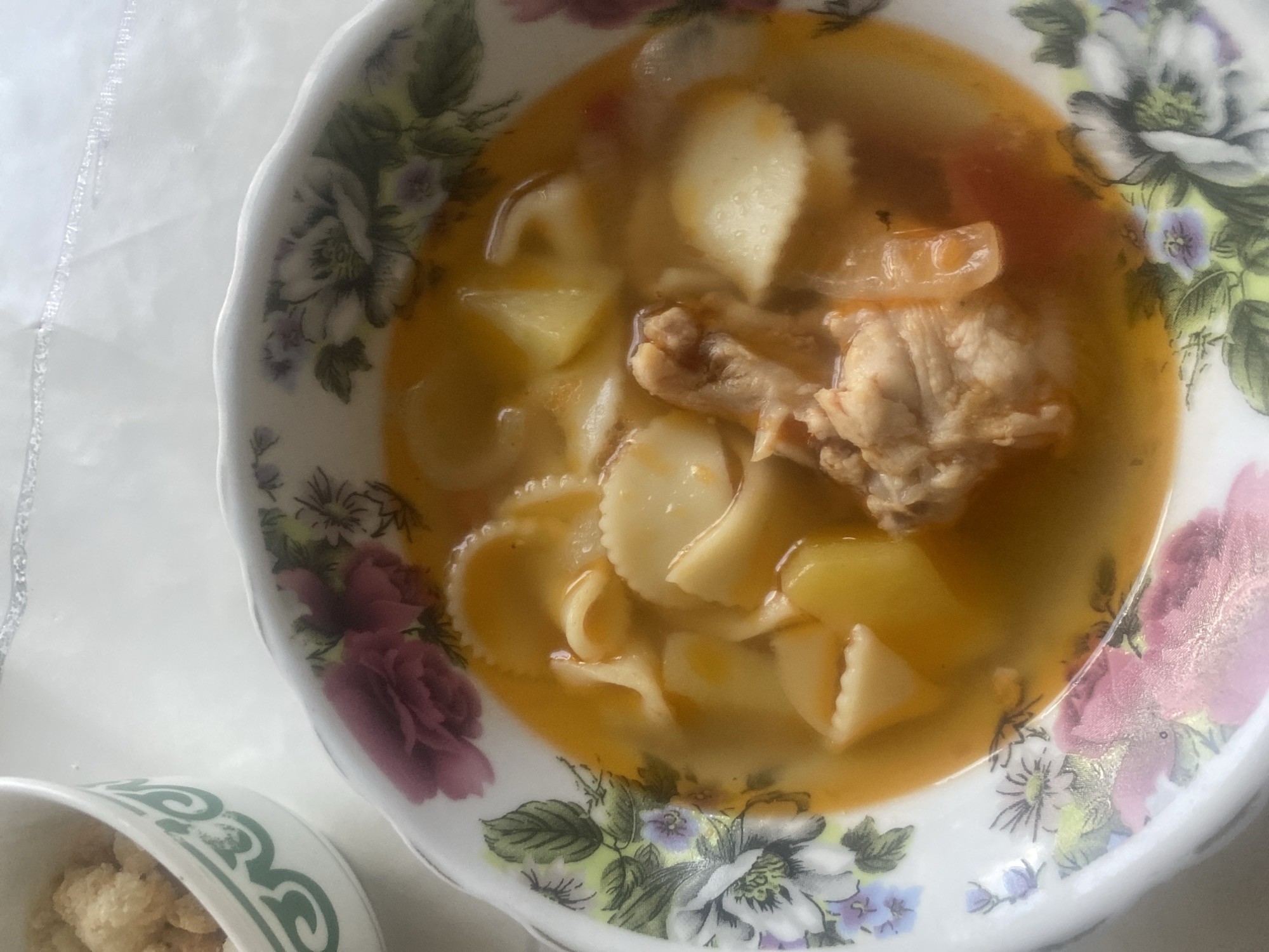 A close-up, overhead shot shows a bowl of soup, likely a traditional Kazakh dish like laghman, with pasta, potatoes, and chicken. The soup is in a decorative floral bowl. To the left of the soup bowl is a small white bowl filled with what appears to be croutons or small bread pieces, also decorated with a green pattern. The background is a white, slightly textured surface, possibly a table. The image is well-lit, with the focus on the food. No people or other elements are visible. The scene is set indoors, likely in a home environment.
