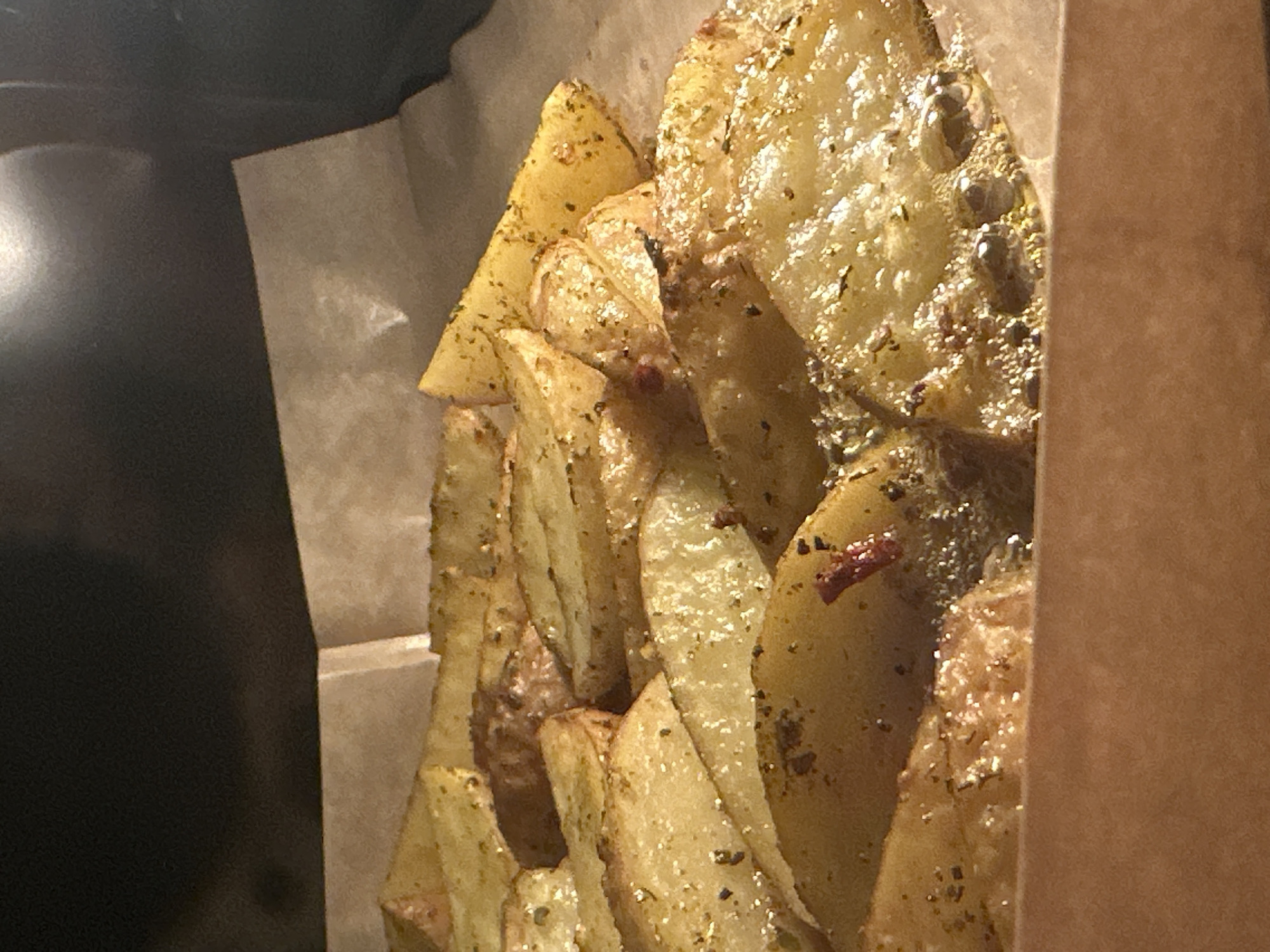 The image displays a close-up, elevated view of numerous seasoned potato wedges resting on a sheet of light brown parchment paper. The paper is contained within what appears to be a light brown cardboard tray or disposable food container, the upper edges of which are visible at the bottom and sides of the frame. The potato wedges exhibit a golden-brown exterior, indicative of cooking, and are generously coated with visible green flakes of dried herbs and smaller dark specks, likely ground pepper or other spices. Some red flakes, possibly chili or paprika, are also discernible among the seasoning. A shimmering liquid, consistent with cooking oil or fat, has pooled around the base of several potato pieces at the bottom of the container. The background above the food and container is uniformly dark and out of focus, suggesting an enclosed space such as an oven or air fryer, with a warm light source illuminating the potatoes from the front or above. No individuals or overt actions are depicted. This scene is observed in Blagoveshchensk, Russia.