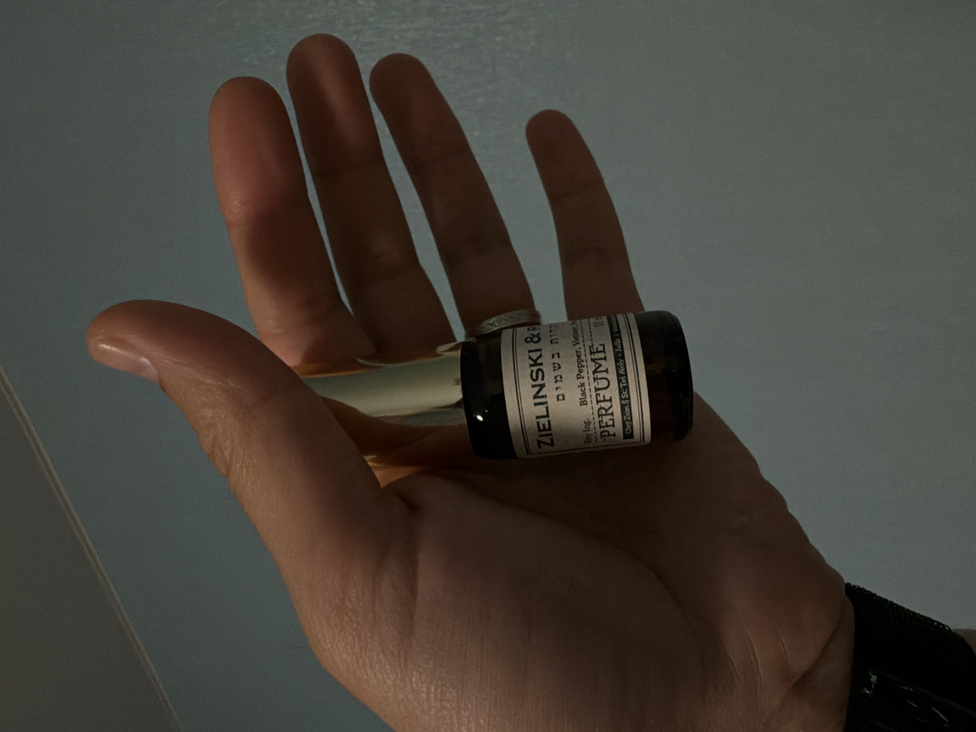 A hand, adorned with a silver ring, holds a small bottle of Zielinski & Rozen perfume. The label indicates the key ingredients as black pepper and vetiver and states "Perfume" in English, along with Hebrew text above the ingredient list. The label also lists the location as Oleg Zion 5 St, Tel Aviv - Jaffa.