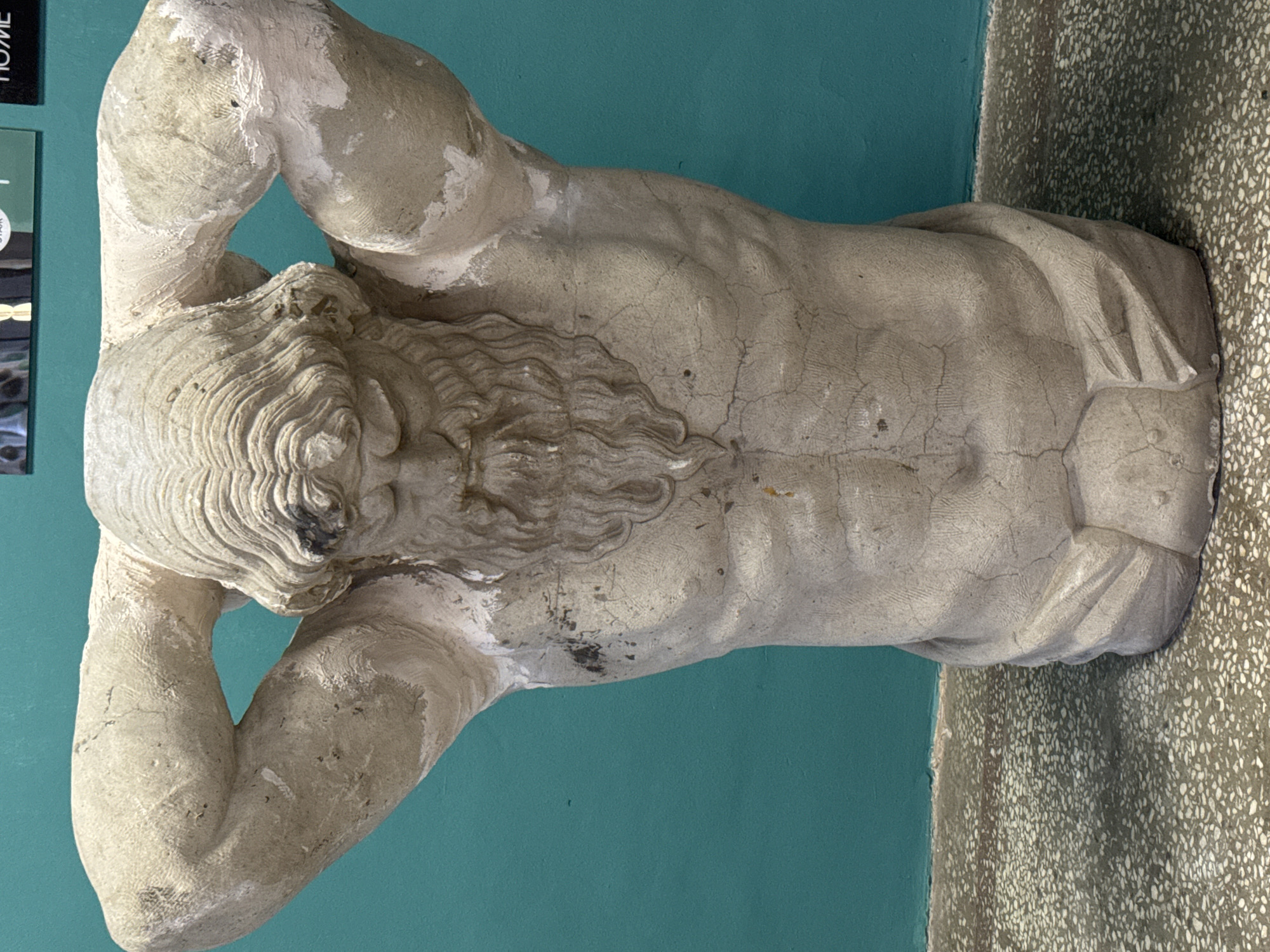 The image shows a male statue with a muscular build. The statue is weathered and appears to be made of stone or plaster. The statue's arms are raised, with the hands placed behind the head. The statue is set against a teal-colored wall and rests on a mottled surface. The statue is a herm, a classical architectural ornament, at the New Hermitage Museum in Saint Petersburg, Russia.