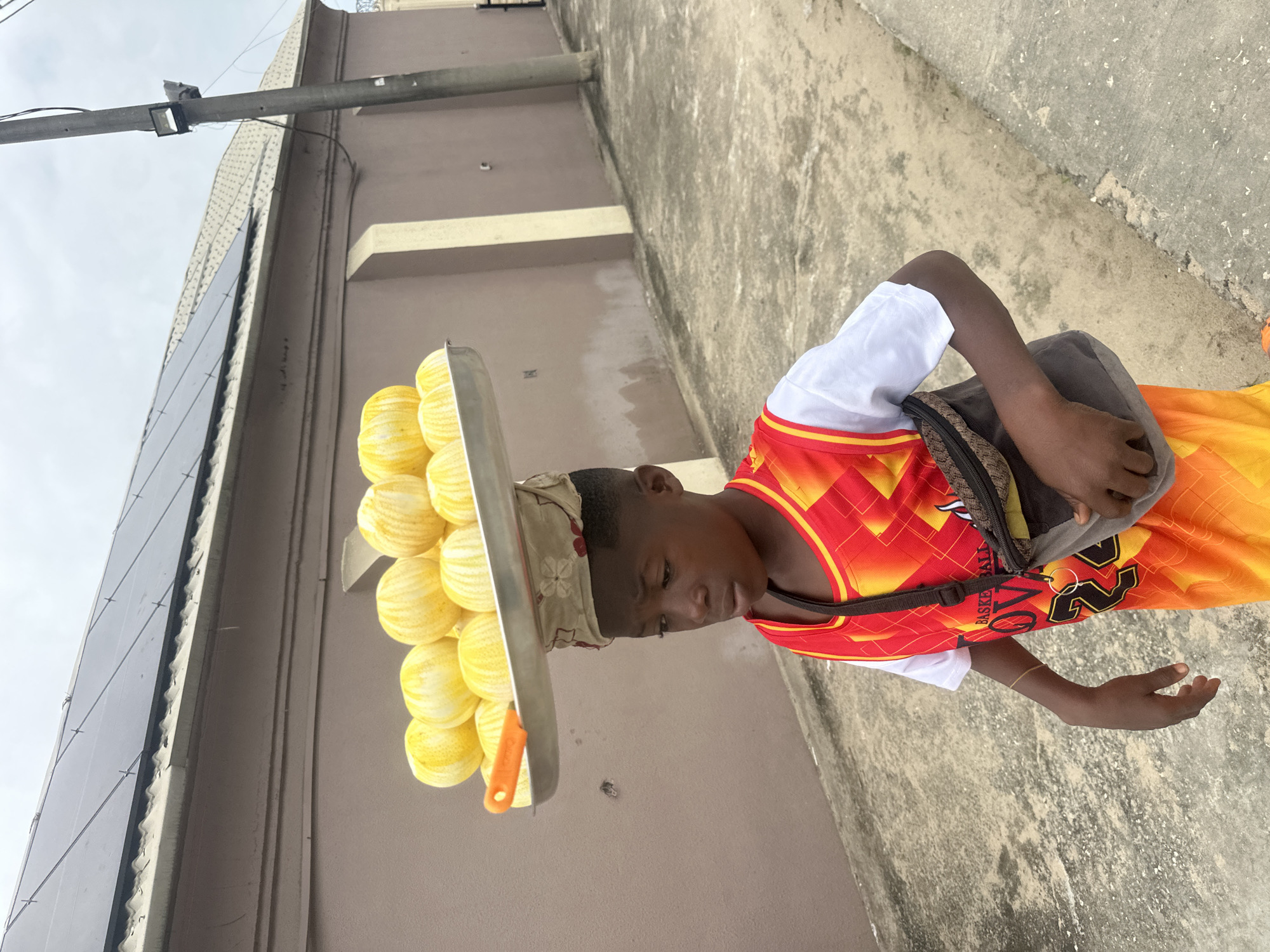 A young boy walks down a paved street in Badagry, Nigeria, carrying a large metal tray of ripe, striped melons balanced on his head. He wears a white t-shirt under a vibrant red and orange basketball jersey. A dark-colored fanny pack is slung across his chest. Behind him, a muted brown building with solar panels on its roof stretches across the background, flanked by a tall gray utility pole. The ground is a textured concrete walkway.