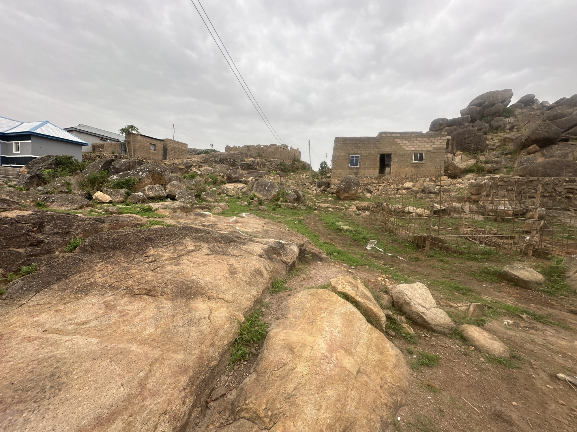 The image depicts a rugged landscape in Jos, Nigeria, characterized by large, smooth, tan-colored rocks dominating the foreground. A dirt path winds through the rocks, leading towards a partially constructed building made of light-colored bricks in the middle ground. The building has visible window and door openings but appears unfinished. A simple, wire fence surrounds a portion of the building.
Further in the background, additional partially constructed or older buildings are visible, interspersed with more rocks and sparse vegetation. A modern, gray house with a blue roof is seen on the left. Overhead, power lines stretch across the cloudy sky. The overall environment suggests a rural or peri-urban setting, where construction and natural rock formations coexist.