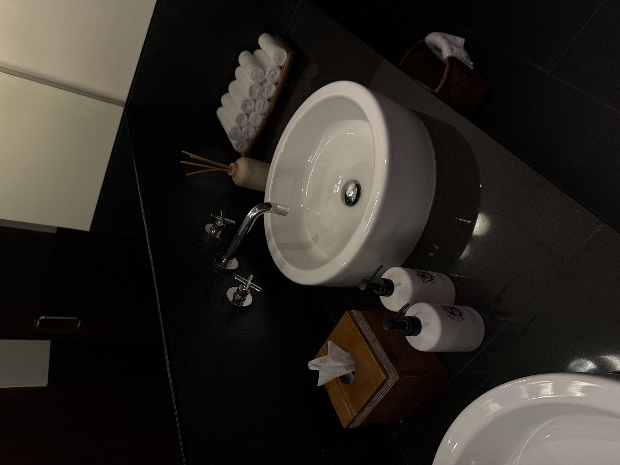 In a dimly lit, modern bathroom, a white ceramic sink is the focal point, with a chrome faucet. To the left of the sink is a wooden tissue box filled with white tissues. Two white pump bottles, likely soap or lotion, stand next to the tissue box. In the background, a wooden tray holds several neatly rolled white towels, and a small vase with thin reeds sits behind them. A dark countertop and black wall dominate the scene, with a reflection in the upper right corner suggesting a mirror and possibly a door. The floor is also dark. The overall impression is one of minimalist luxury.