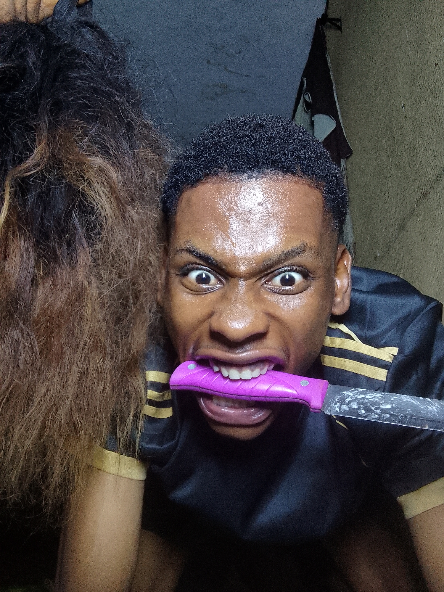 A close-up, harshly lit photograph shows a young Black man with short, dark hair and wide eyes, holding a knife in his mouth. The knife has a purple handle and a silver blade. The man is wearing a black athletic shirt with yellow stripes on the sleeves. To the left of the man is a person with long brown hair. The background is a dimly lit interior with textured walls and an unidentified object hanging on the wall in the upper right corner. The location appears to be indoors, but the specific location is unidentified. The man's expression is intense, with his teeth bared.