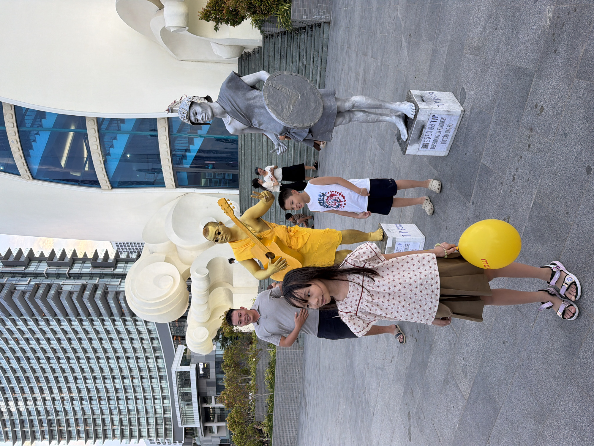 In Nha Trang Ward, Vietnam, a scene unfolds with two street performers dressed as statues, one golden and the other silver, positioned on raised platforms. The golden performer, adorned in a yellow robe, strums a guitar, while the silver performer, clad in a tunic and helmet, holds a round shield. In the foreground, a young girl with long dark hair, wearing a patterned blouse and brown skirt, clutches a bright yellow balloon. To her left, a man in a grey polo shirt and dark shorts stands watching. Behind them, a young boy in a white t-shirt and dark shorts observes the silver statue. In the background, a tall, modern building with numerous balconies dominates the skyline, contrasting with the white, sculptural architecture of a nearby structure. The ground is paved with grey tiles, and a few other people are visible in the distance.