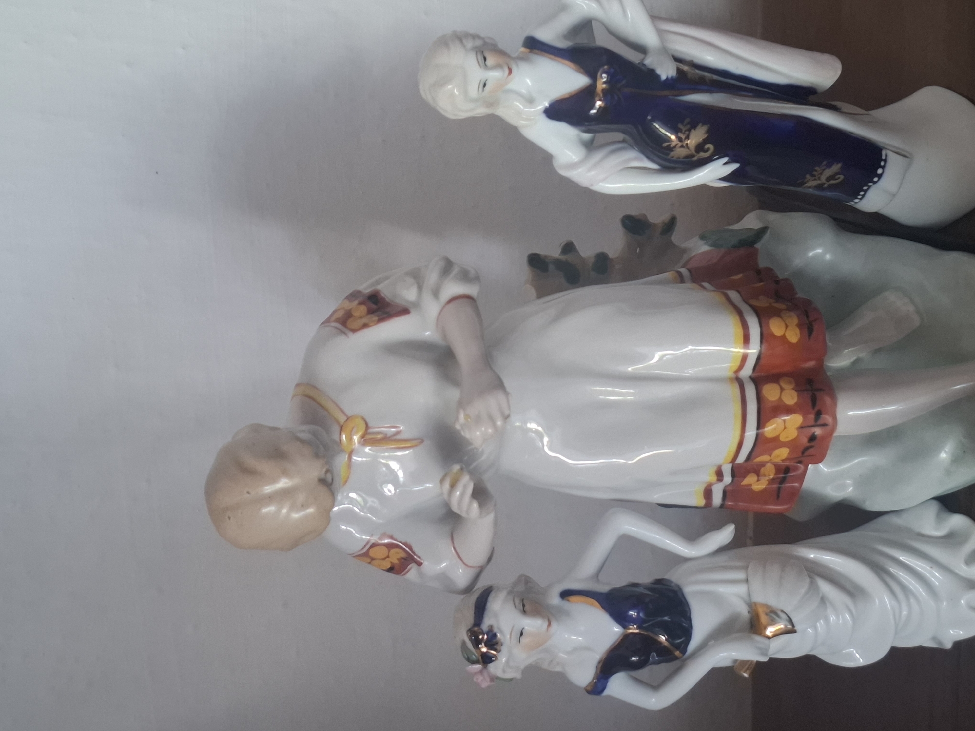 The image shows three porcelain figurines arranged on a surface, likely a shelf or table. The figurines are of women in detailed dresses. The central figure is larger and appears to be wearing a white dress with a red and yellow floral border. She is bending forward, with her hands near her waist. To the left, a smaller figurine is dressed in a white gown with a dark blue bodice adorned with gold accents, and a pink flower in her hair. To the right, another figurine wears a similar white gown with a dark blue dress featuring gold leaf patterns. The background is a plain, light-colored wall. There is no discernible location in the city of Almaty, Kazakhstan, indicated in the image.