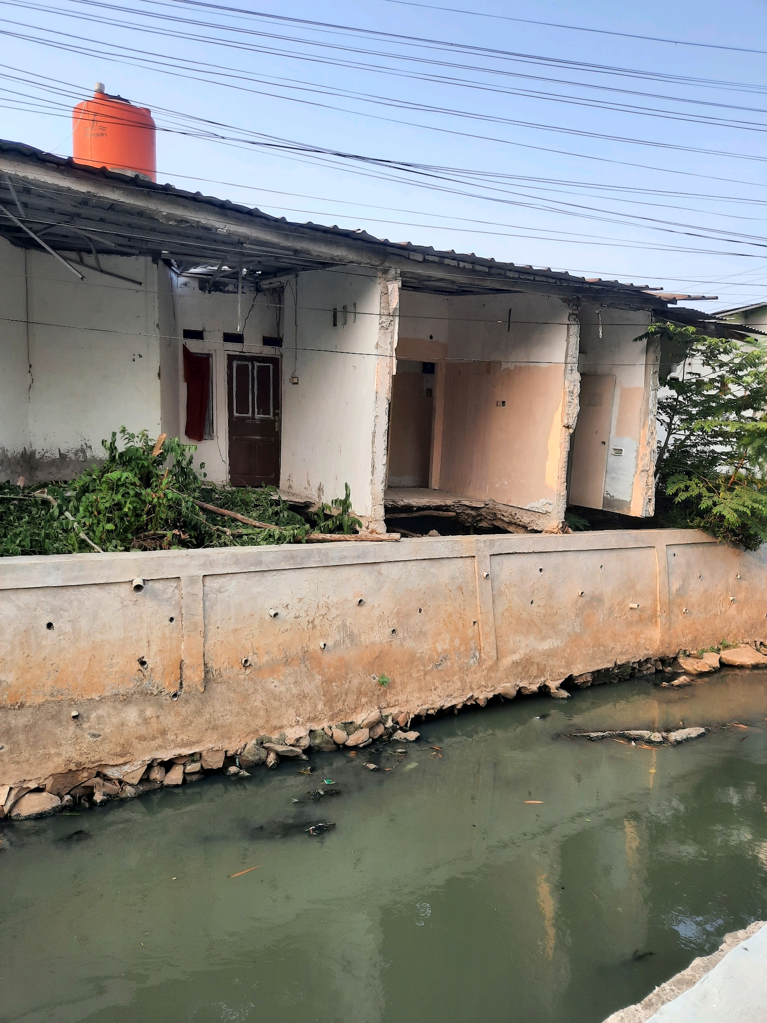 The image shows a section of a canal in East Jakarta, Indonesia, with a damaged building on its bank. The building appears to be in a state of disrepair, with its roof partially collapsed and walls crumbling. Debris, including branches and leaves, is scattered around the base of the building and on the canal bank. The canal itself contains murky, greenish water with some debris floating on the surface. The scene is under a clear sky with power lines visible above the building.