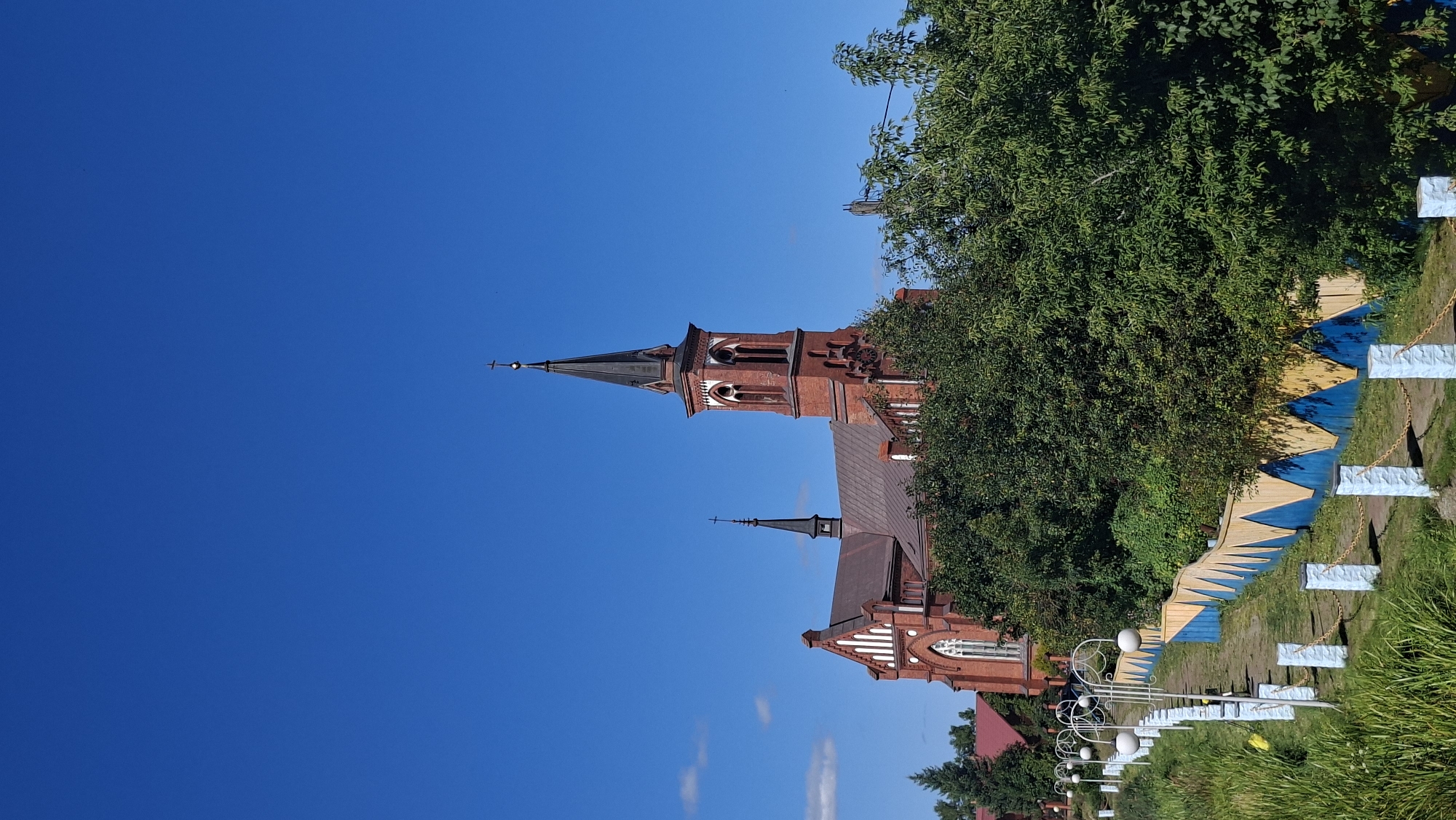 The image shows a brick church with a tall steeple, located in Pastavy, Belarus. The church is surrounded by lush green trees and foliage. A pathway with decorative fencing and lampposts leads up to the church. The sky is a clear, bright blue with a few scattered white clouds. The overall scene depicts a peaceful and picturesque religious site under a sunny sky.