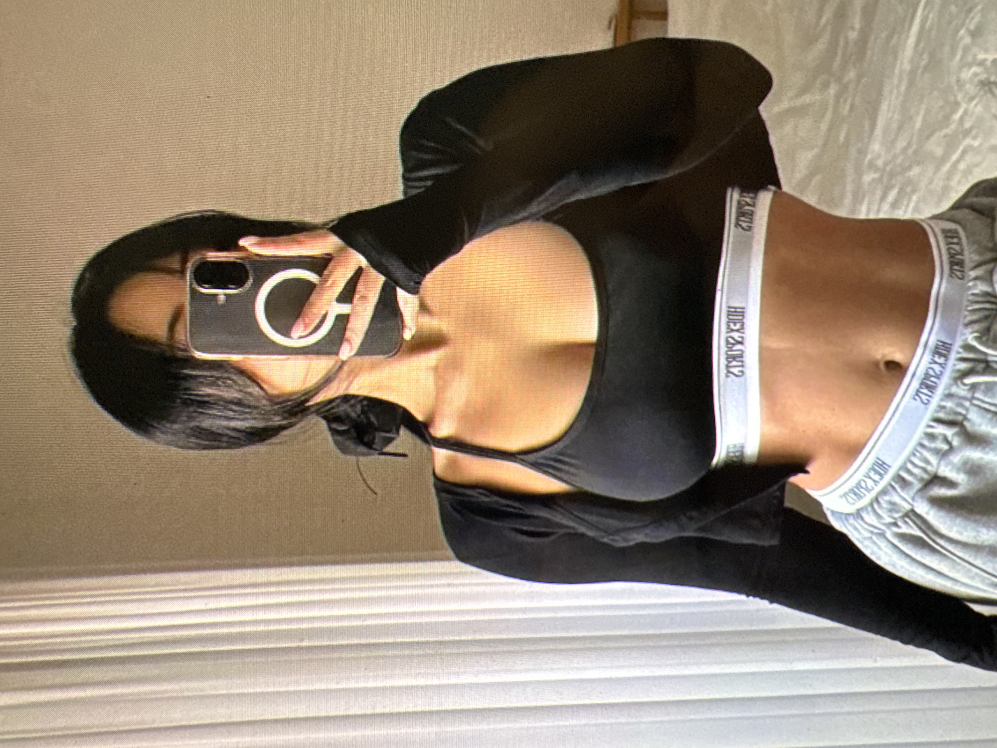 A person with dark hair is taking a mirror selfie in what appears to be a bedroom. They are wearing a black sports bra, a long-sleeved black crop top, and grey sweatpants. The person is holding up a smartphone to take the picture, obscuring their face. The background shows a white vertical blinds, a bed, and a wooden headboard. The image lacks any identifiable landmarks or details that would indicate its location in Castelo Branco, Portugal.