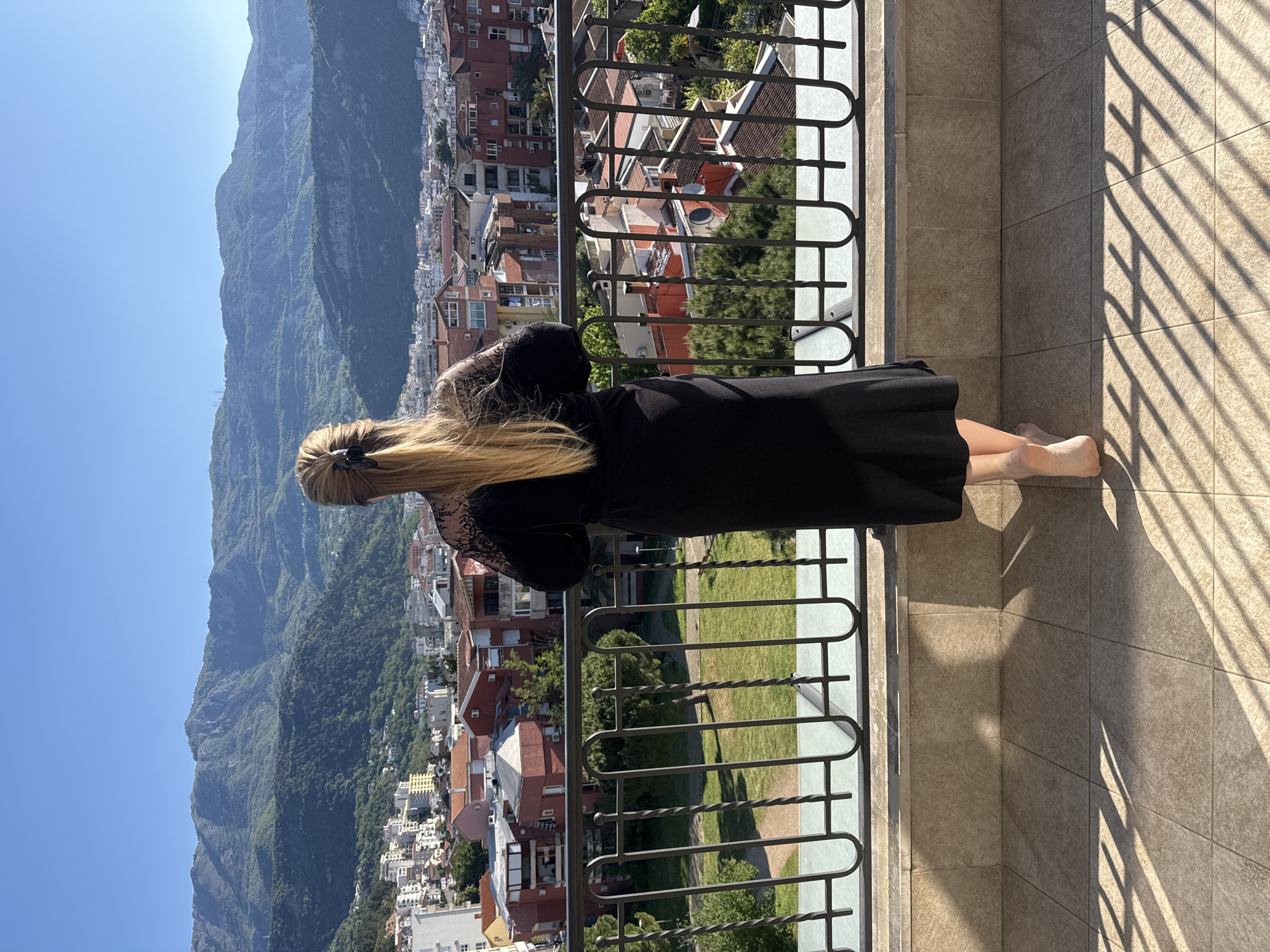 The image shows a woman standing on a balcony overlooking a cityscape with a mountain range in the background. The woman has long blonde hair and is wearing a black dress. She is facing away from the camera, looking out at the view. The balcony has a metal railing. The cityscape consists of buildings with red roofs. The sky is blue. Based on the architecture and mountainous landscape, this is likely Budva, Montenegro.