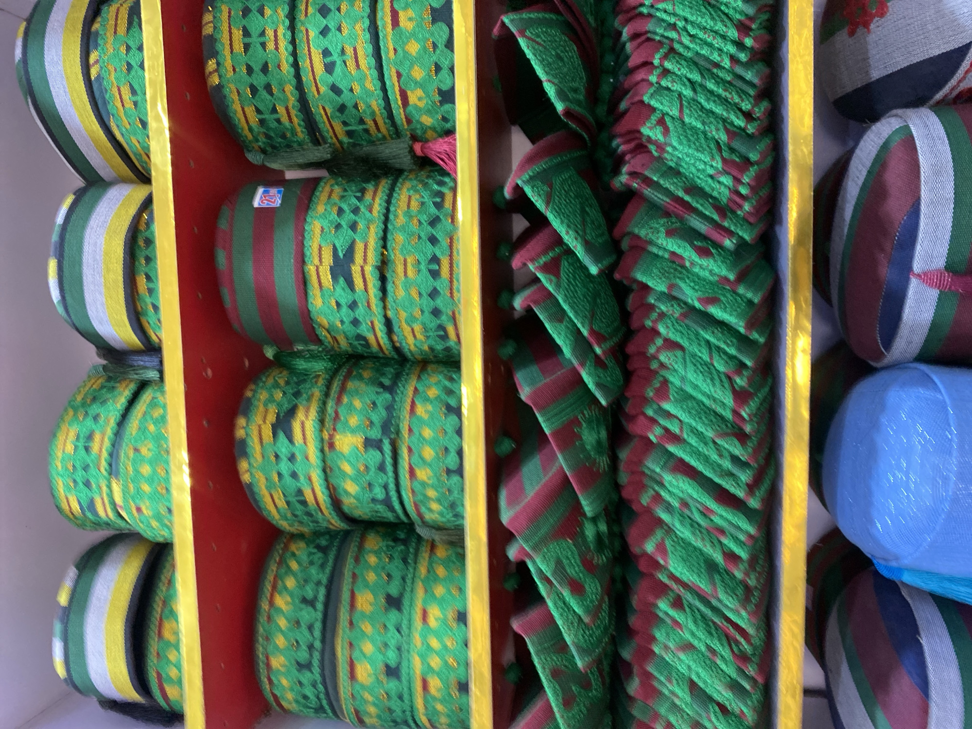 The image shows shelves of colorful, cylindrical objects. The objects appear to be woven or embroidered fabric, possibly hats or caps. Some are stacked and patterned with green, yellow, and red. Others are arranged vertically, showing green and red stripes or triangular patterns. The shelves are a combination of yellow and red. The image was taken in the city of Bukuru, Nigeria.