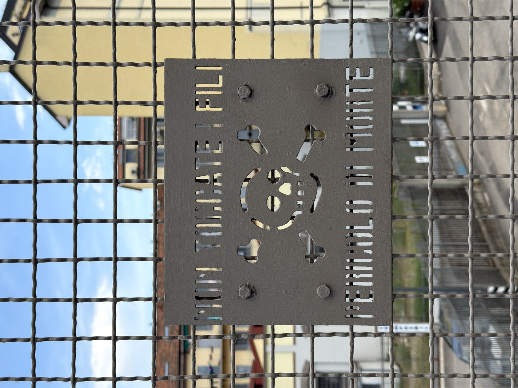 A grey, square sign with a skull and crossbones symbol is affixed to a metal mesh fence. The sign is bolted to the fence at its four corners. The left side of the sign has the Italian words "NON TOCCATE I FILI" cut out in vertical lettering, while the right side has "PERICOLO DI MORTE" similarly cut out. Through the cut-out portions of the sign, parts of buildings, sky, and possibly other street elements are visible. The fence itself has a grid-like pattern. The location is Milan, Italy.