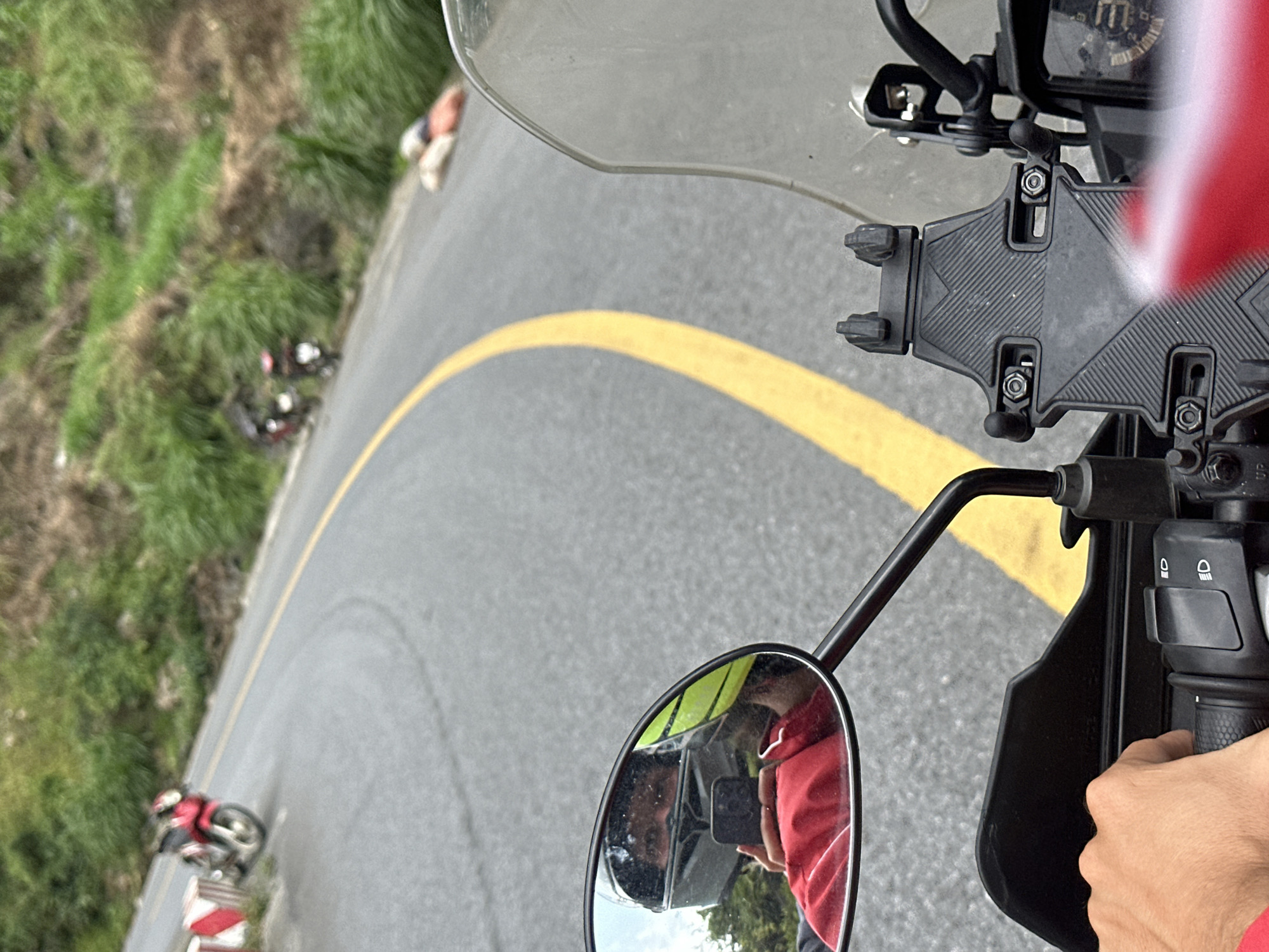 The image provides a first-person perspective from a motorcycle, looking down a winding asphalt road in what appears to be Minh Sơn Commune, Vietnam. The main subject in the immediate foreground consists of parts of a motorcycle: a clear windshield, dashboard with faint gauges, a black phone mount (currently empty), and the right handlebar with control switches. The rider's right hand, wearing a red sleeve, grips the handlebar.
Reflected in the circular rearview mirror on the left, the person taking the photo is visible, wearing a red top and holding a smartphone to capture the image. The road ahead curves sharply to the left, marked by a prominent, continuous double yellow line indicating a no-passing zone. To the left of the road are lush green grassy slopes and hills.
Further down the curved road, another red motorcycle, possibly a touring or sport bike, is visible on the left side near a red and white barrier. Near this motorcycle, a person is standing or walking alongside the road. The overall setting suggests a scenic, hilly, or mountainous area. The lighting indicates it is daytime, likely a clear day. No specific emotions are discernible, but the scene conveys a sense of a journey or a stop during a ride. Visible text on the motorcycle dashboard is too indistinct to read, apart from a possible "UP" or symbol.