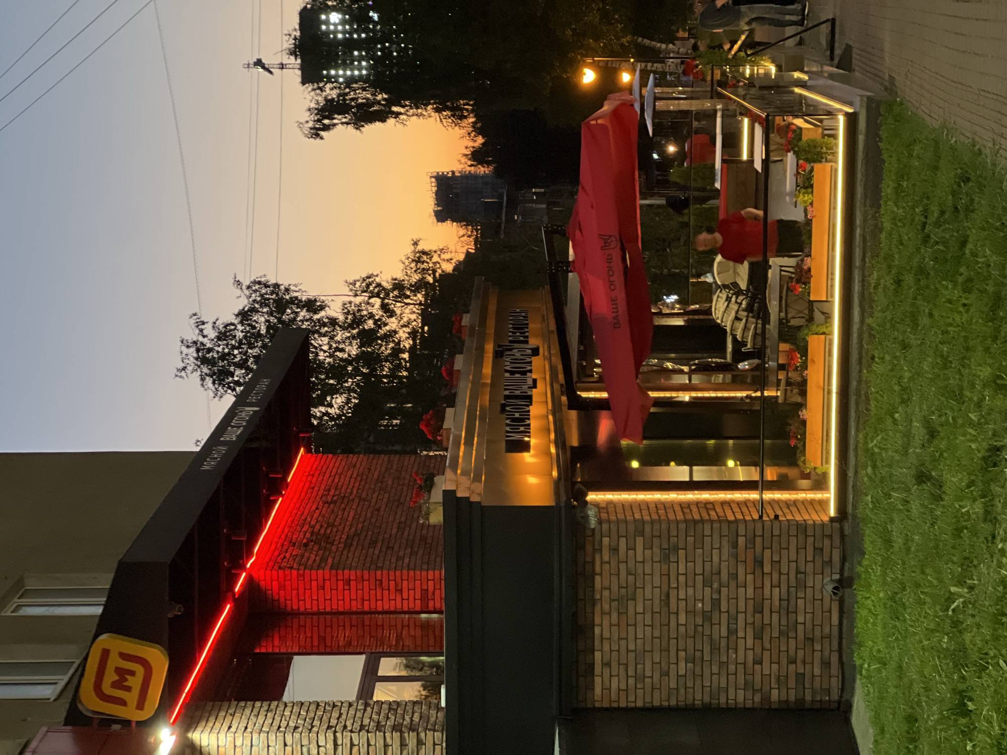 The image captures a restaurant facade in Izhevsk, Russia, at dusk. The establishment, identified by a yellow "M" logo and the text "МЯСНОЙ ВАШЕ ОГОНЬ РЕСТОРАН" (Miasnoy Vashe Ogonyok Restoran - Meat Your Fire Restaurant), is adorned with bright red neon lights along its overhang and warm orange string lights illuminating the ground level. The building's exterior is constructed with brick. A red umbrella covers an outdoor seating area where a man in a red shirt is visible. In the background, trees and city buildings, including a construction site, are silhouetted against an orange and purple gradient sky. The scene is set beside a grassy area and a paved walkway.