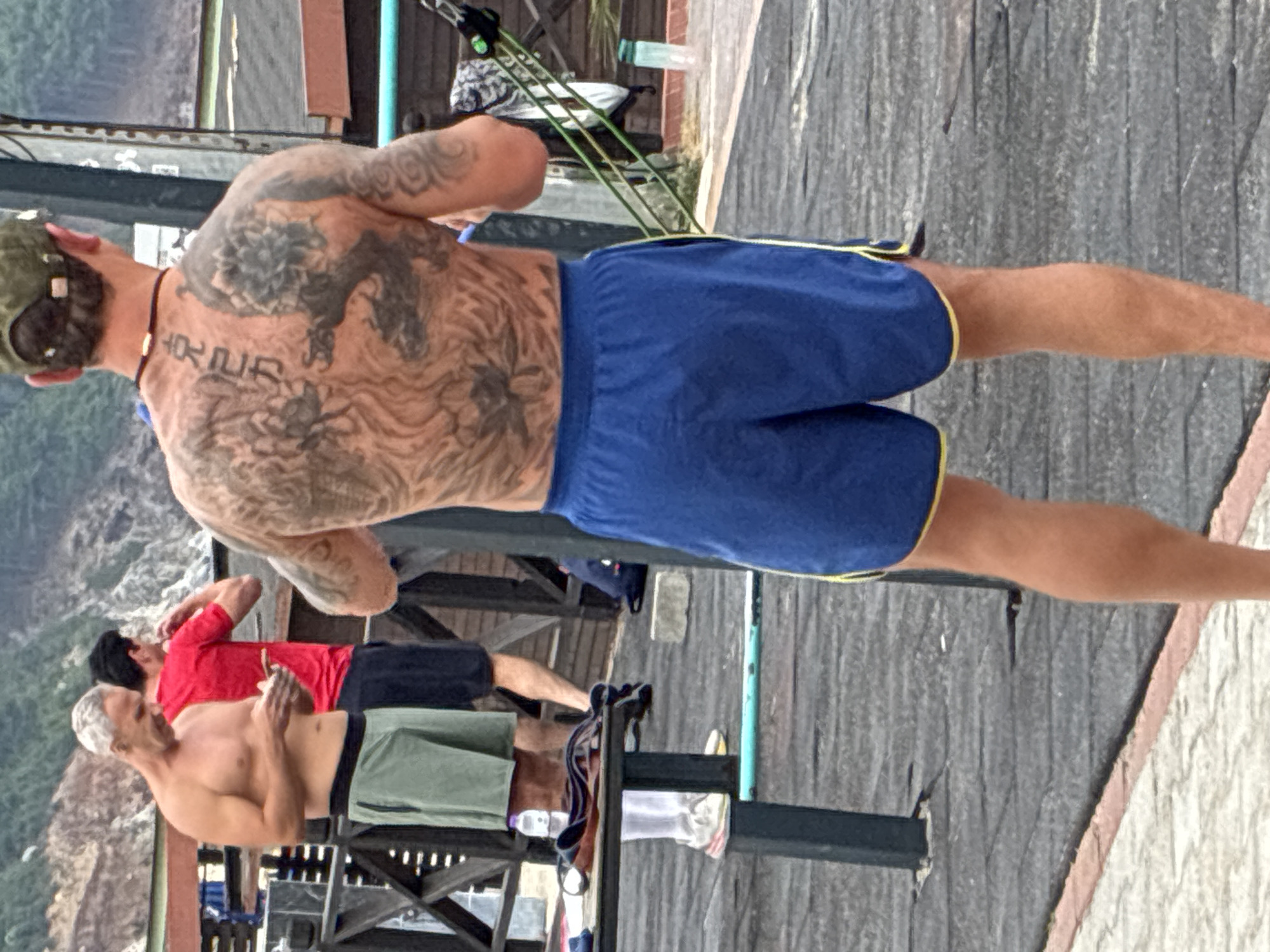 Two adult males are visible outdoors, with a third male partially in frame. The primary subject, a male with a heavily tattooed back, stands facing away from the camera, wearing a green baseball cap and blue athletic shorts with yellow trim. He has a necklace around his neck and appears to be engaged in an activity involving resistance bands. The second visible male, to the left and further back, is shirtless with graying hair and is wearing light green athletic shorts. He is looking down at his phone. The third male, behind and to the right of the primary subject, wears a red t-shirt and dark shorts and is looking towards his left. The background consists of wooden structures, foliage, and a hillside. The ground is a dark, textured surface, possibly a wooden deck or asphalt. There are various objects scattered around, including a water bottle, a towel, and a gray concrete block. The lighting suggests an overcast day. The scene appears to be an outdoor workout or recreational area.