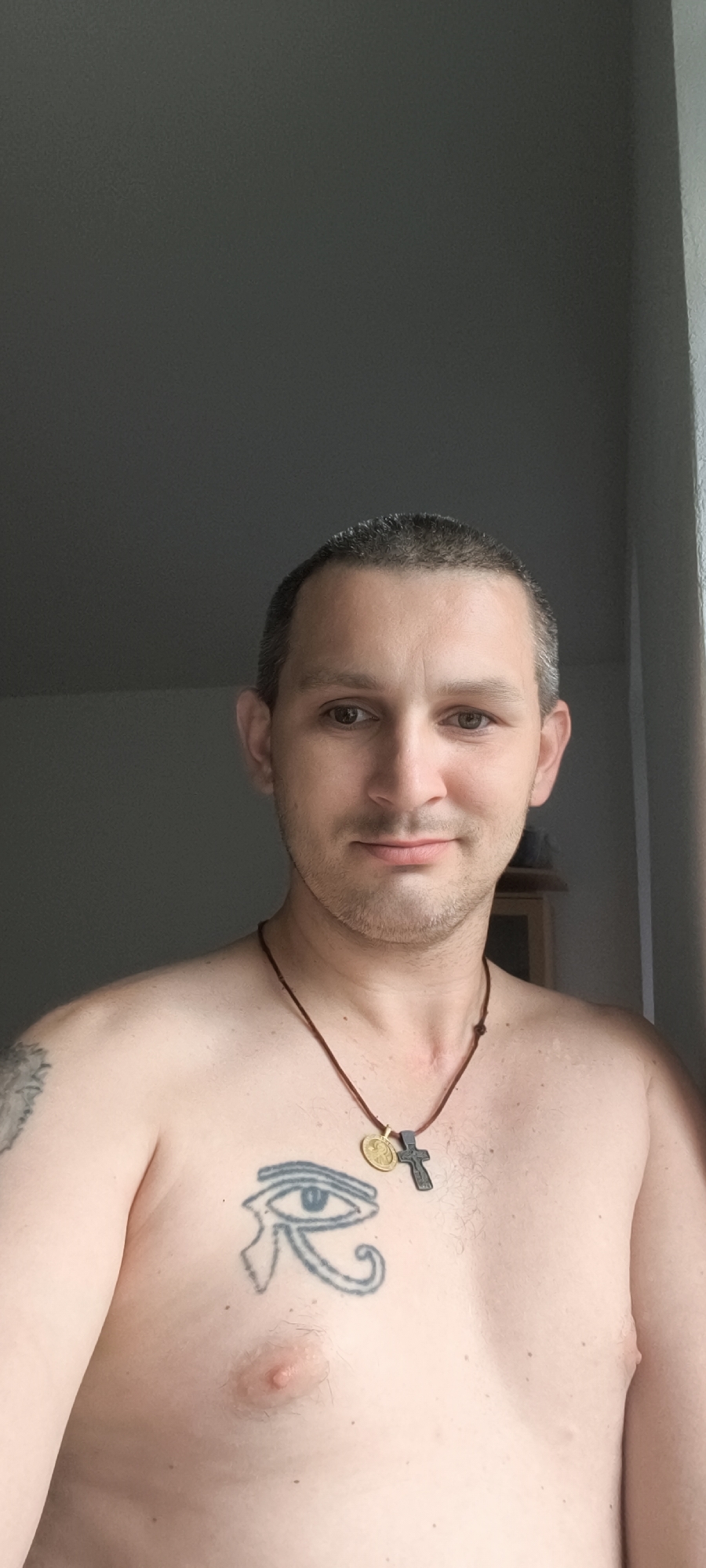 A person is taking a chest-up selfie in what appears to be an indoor setting with natural light from the right. The individual has short, dark hair, brown eyes, and is not wearing a shirt. They have a tattoo of the Eye of Horus on their chest and a tattoo on their left shoulder/upper arm that is partially visible. They are wearing a brown cord necklace with two pendants: a small gold-colored circular pendant and a dark cross-shaped pendant. The background is out of focus and appears to be a wall, with a hint of a wooden cabinet or shelf visible on the right. The overall lighting suggests it is daytime. There is no information in the image to indicate the location in Wolgast, Germany.