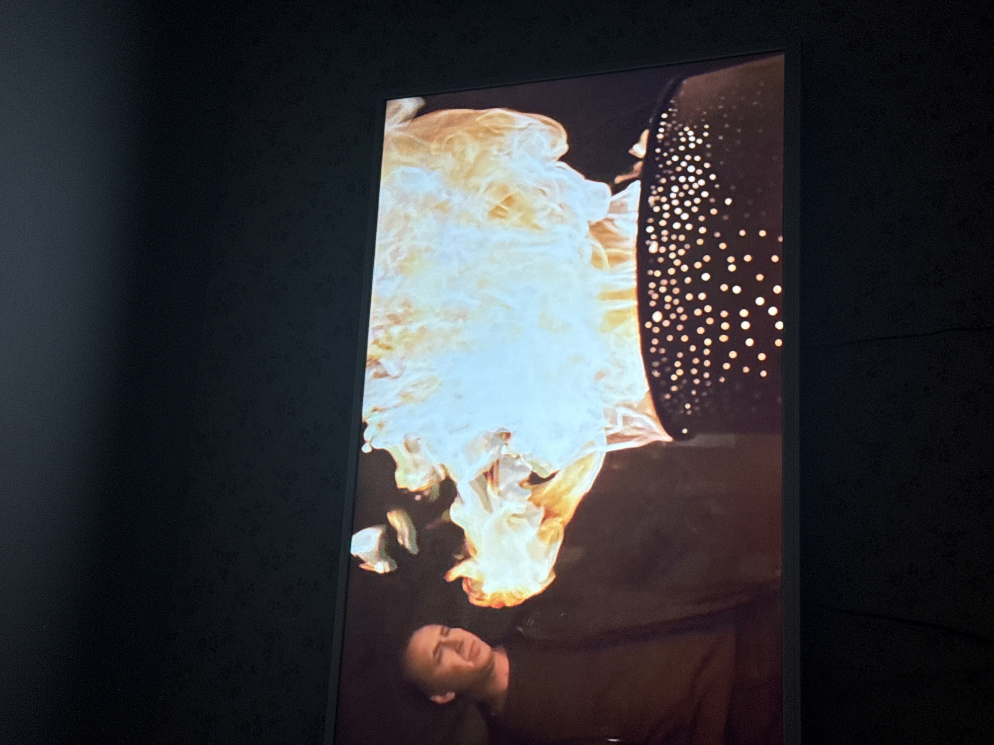 A television screen, located indoors in an unknown location in Mytishchi, Russia, displays a scene from a video. The scene shows a person lying down, facing the camera. Above the person is a column of bright orange and white flames. Adjacent to the flames on the right side is a dark, vertical figure with a pattern of evenly spaced white dots. The screen is surrounded by a dark, textured wall.
