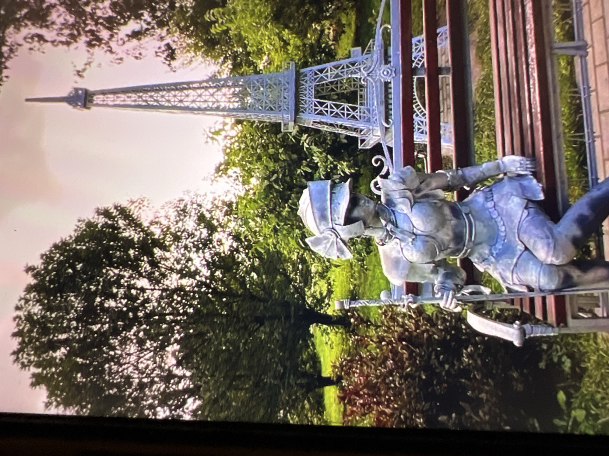 The image shows a statue of a female figure in armor sitting on a park bench. The statue is a metallic gray color and depicts the figure wearing a helmet with a large bow on top, a chest plate, arm guards, and a skirt. She is holding a long staff-like object in her right hand. Behind the statue, there is a miniature replica of the Eiffel Tower. The background consists of lush green trees and grass, suggesting an outdoor park setting. The lighting indicates it is daytime. The statue is located in the "Park of the Great Patriotic War" in Minsk, Belarus.