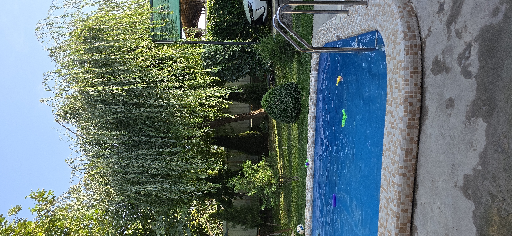 The image shows a swimming pool in Yusuphana, Uzbekistan, with a large weeping willow tree in the background. The pool is filled with bright blue water and has a tiled edge. Several colorful toys are floating in the pool, including a yellow duck, two green water guns, and a purple float. A soccer ball is visible on the grass near the pool. The area around the pool is a well-maintained garden with manicured bushes, trees, and a fence. A white car is parked to the right of the frame, partially obscured by vegetation. The sky is clear and blue, indicating fair weather.