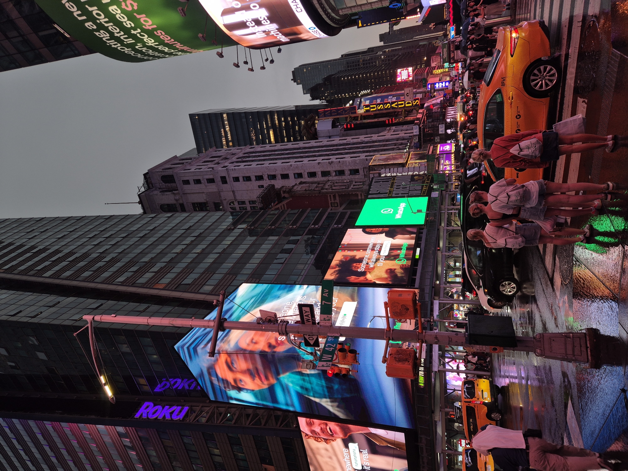 This is a street-level, eye-level, medium shot of Times Square in New York City at dusk. The sky is overcast, and the street is wet and reflective, indicating recent rain. Large, bright digital billboards dominate the background, displaying advertisements and graphics. On the left, a tall building features a prominent purple "Roku" logo illuminated neon. Below this, a massive screen shows a close-up of two faces. A traffic light and street signs are visible in the center, with "W 42 ST" and "7 AV" clearly marked. Several people are present in the scene, some holding umbrellas, suggesting continued light rain or drizzle. On the right side of the image, a yellow taxi is parked near a crosswalk where three young women are standing, appearing to observe the surroundings. More people and vehicles are visible further down the street, contributing to the bustling atmosphere of Times Square.