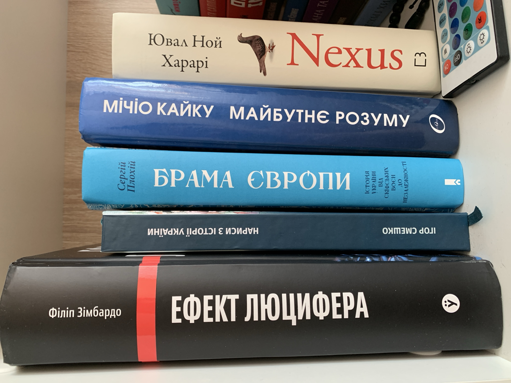 A close-up, eye-level, vertical shot shows a bookshelf with six books. The books are arranged from left to right. The leftmost book is black with white text that reads "Філлін Зімбардо" at the top and "ЕФЕКТ ЛЮЦИФЕРА" vertically. To the right of this book is a dark blue book with white text that reads "НАРИСИ З ІСТОРІЇ УКРАЇНИ" vertically. Next to that is a bright blue book with white text that reads "Сергій Плохій" at the top and "БРАМА ЄВРОПИ" vertically. To its right is a dark blue book with white text that reads "Мічіо Кайку" at the top and "МАЙБУТНЄ РОЗУМУ" vertically. The rightmost visible book has a cream-colored cover with red text that reads "Ювал Ной Харарі" at the top and "Nexus" vertically, with a small illustration of a bird. Behind these books, other books are partially visible. A black remote control with colorful buttons is on the shelf in the foreground on the right. The background shows a light-colored wooden shelf. The image appears to be taken indoors in Ternopil, Ukraine.