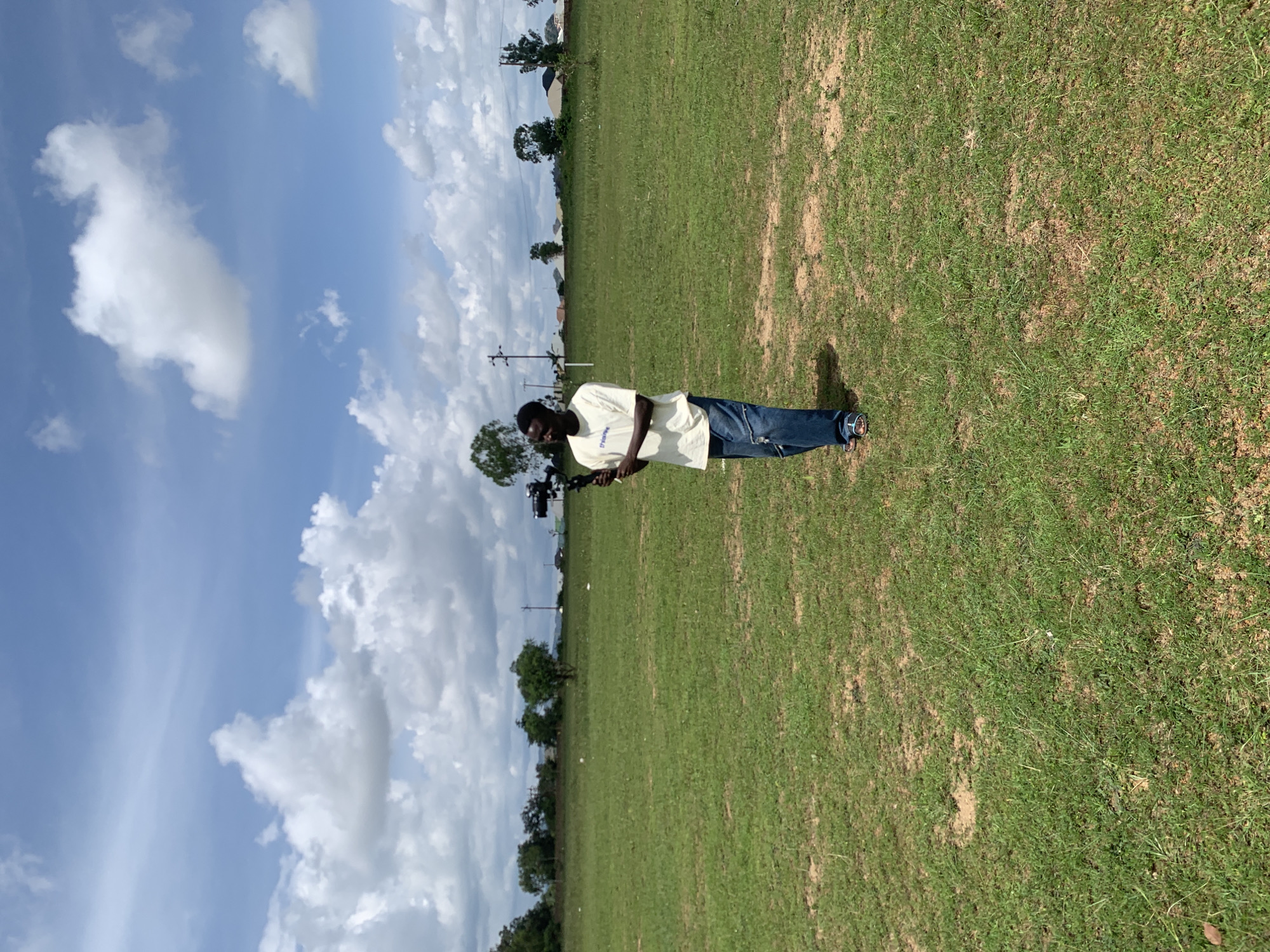 A young Black male stands in a grassy field, holding a professional camera rig with a gimbal. He is wearing a light-colored t-shirt, loose-fitting blue jeans, and sandals. The field is expansive and appears to be in an open, undeveloped area. In the background, there is a line of trees and some scattered buildings, suggesting a semi-urban or rural setting. The sky is blue with scattered white clouds, indicative of a bright, possibly hot day. The scene is likely located in Jos, Nigeria, given the geographical context. The subject appears to be in the process of filming or preparing to film.