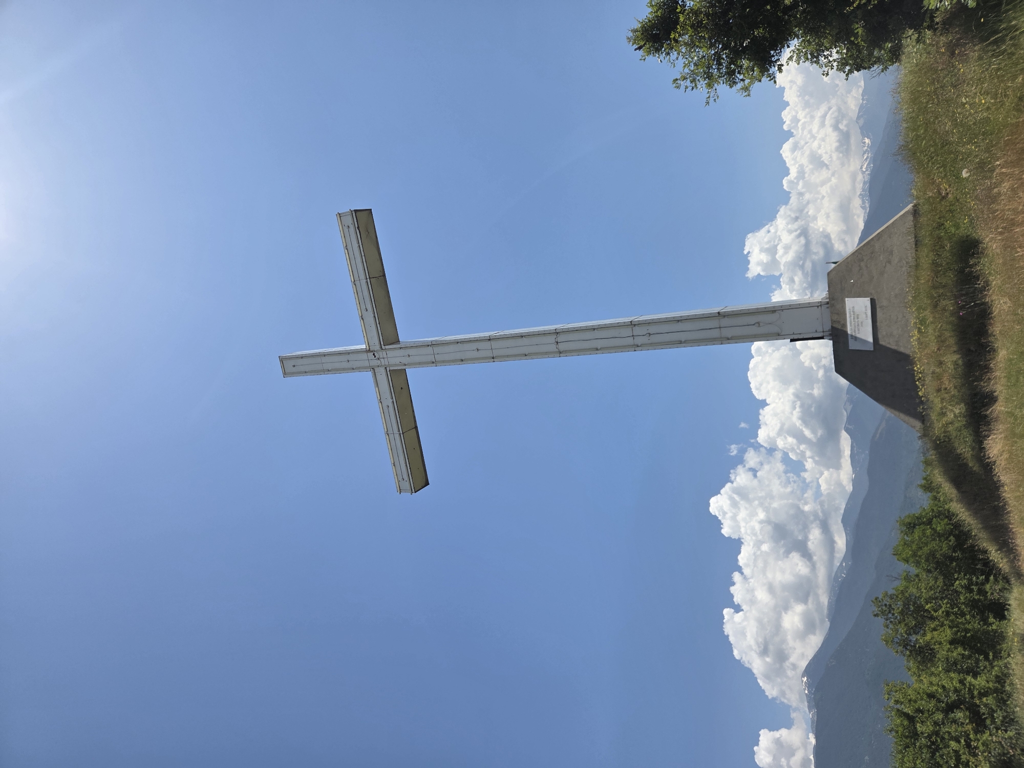 The image captures a large, prominent white cross monument situated on a grassy hill under a clear, bright blue sky with scattered white cumulus clouds, in Brezhani, Bulgaria. The cross, appearing to be constructed from light-colored metal or a similar material, features a box-like structure with visible panel lines. It stands vertically with a horizontal crossbeam. The base of the cross is a trapezoidal concrete structure, broader at the bottom. A rectangular plaque, bearing an inscription and the year '2006', is affixed to the left side of the cross's base. A sun flare is visible in the upper right quadrant of the sky, indicating a bright, sunny day. In the background, multiple ranges of mountains extend across the horizon, with several distant peaks exhibiting patches of snow. The lower slopes of these mountains are covered in dense green foliage. The immediate foreground consists of a grassy slope with a mix of dry, straw-colored grass, greener patches, and small wildflowers. Green trees and bushes are visible on the left and right sides of the frame, flanking the cross. No individuals or specific actions are depicted.