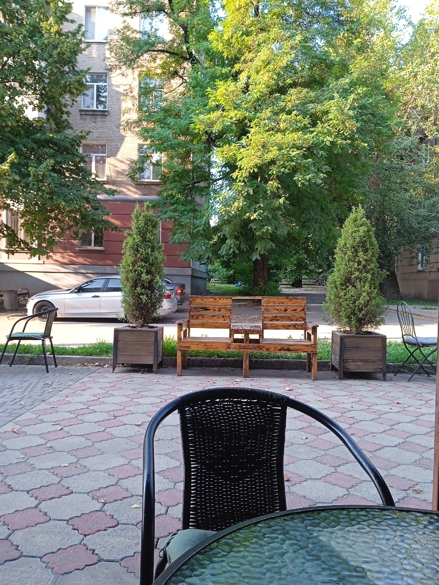 An outdoor seating area is depicted in front of a multi-story brick building. In the foreground, a black woven chair with a glass-topped table is partially visible. Behind this, a wooden bench is situated with a square planter box containing a tall, slender evergreen tree on its right. To the left of the bench, another square planter box holds a similar evergreen tree. A dark metal chair with a seat is visible further to the left, and another similar chair is to the far right. A silver sedan is parked on a paved area behind the planters. The ground is paved with interlocking geometric tiles in shades of gray and pink. Lush green trees provide shade over the scene.