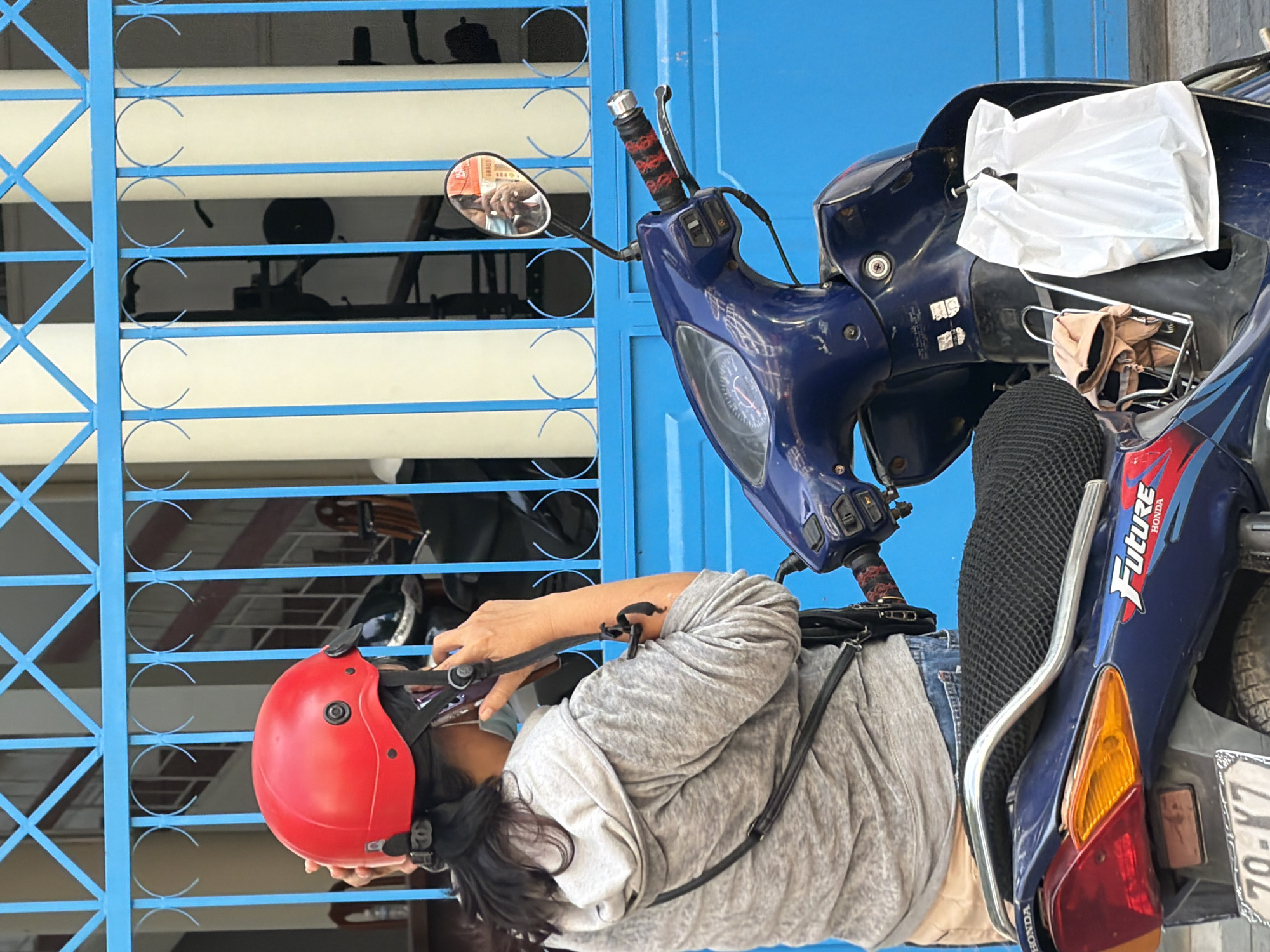 In Nha Trang, Vietnam, a woman wearing a red helmet and a grey hooded jacket is seen from behind, talking on her smartphone. She is standing next to a blue scooter, which has a white plastic bag hanging from its front. The background features a blue metal gate and light-colored pillars, suggesting an outdoor urban setting. The woman's face is partially obscured by her hand and the phone.