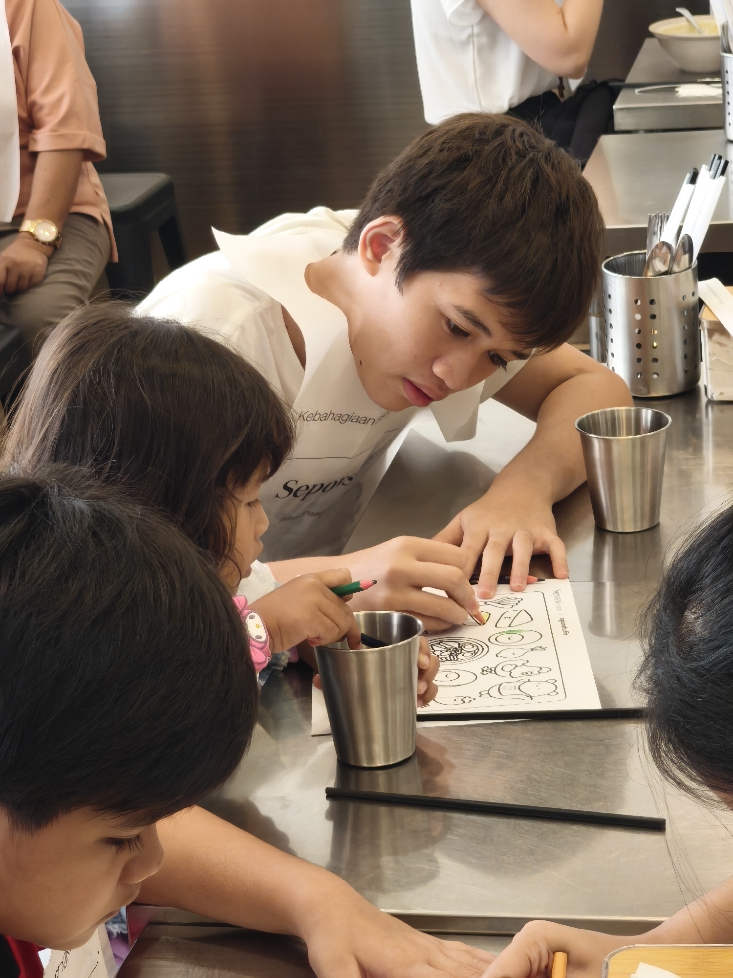 In South Jakarta, Indonesia, a group of children and an adult are engaged in an art activity at a metal table. A young boy, with dark hair and wearing a white t-shirt, leans over a coloring page, his right hand holding a pencil and coloring an image on the page. To his left, a young girl, also with dark hair, appears to be drawing or coloring as well, holding a green pencil. In the foreground, another child with dark hair is partially visible, leaning towards the table. An adult, wearing a peach-colored shirt and beige pants, is seated on a stool in the background, observing the activity. The environment suggests an indoor setting, possibly a cafe or community space, with a cutlery holder containing spoons and chopsticks visible on the table. The overall scene depicts a collaborative and focused artistic endeavor among the individuals.