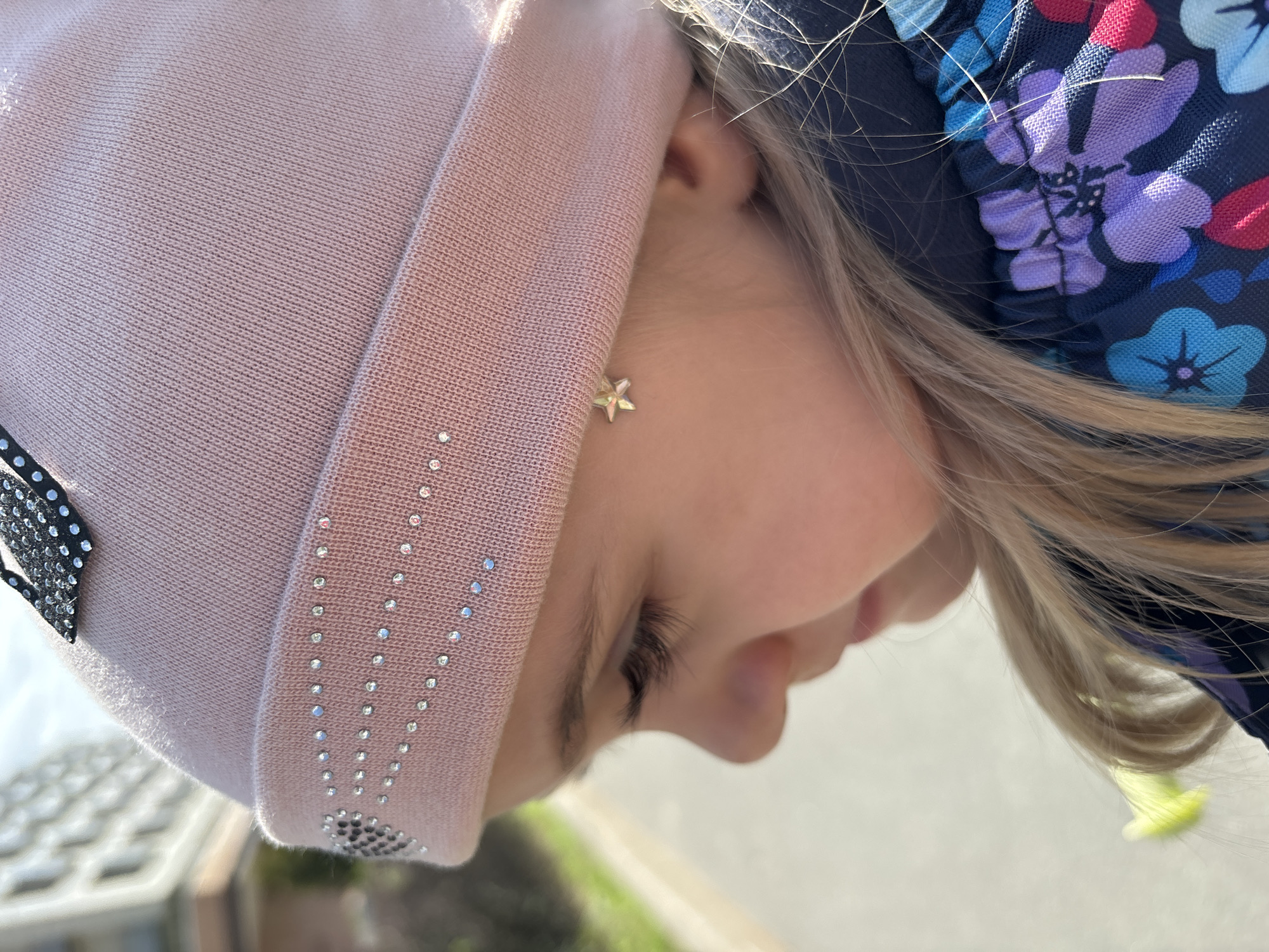 A close-up, eye-level shot depicts a young child with blonde hair, wearing a light pink knitted hat adorned with small rhinestones and a flower-patterned navy blue hat on top. The child's face is in profile, showing a small star-shaped earring. The background suggests an outdoor setting, possibly in Saint Petersburg, Russia, with elements of pavement and greenery visible. The child's downward gaze and the tilt of the frame suggest the child may be looking at something below.