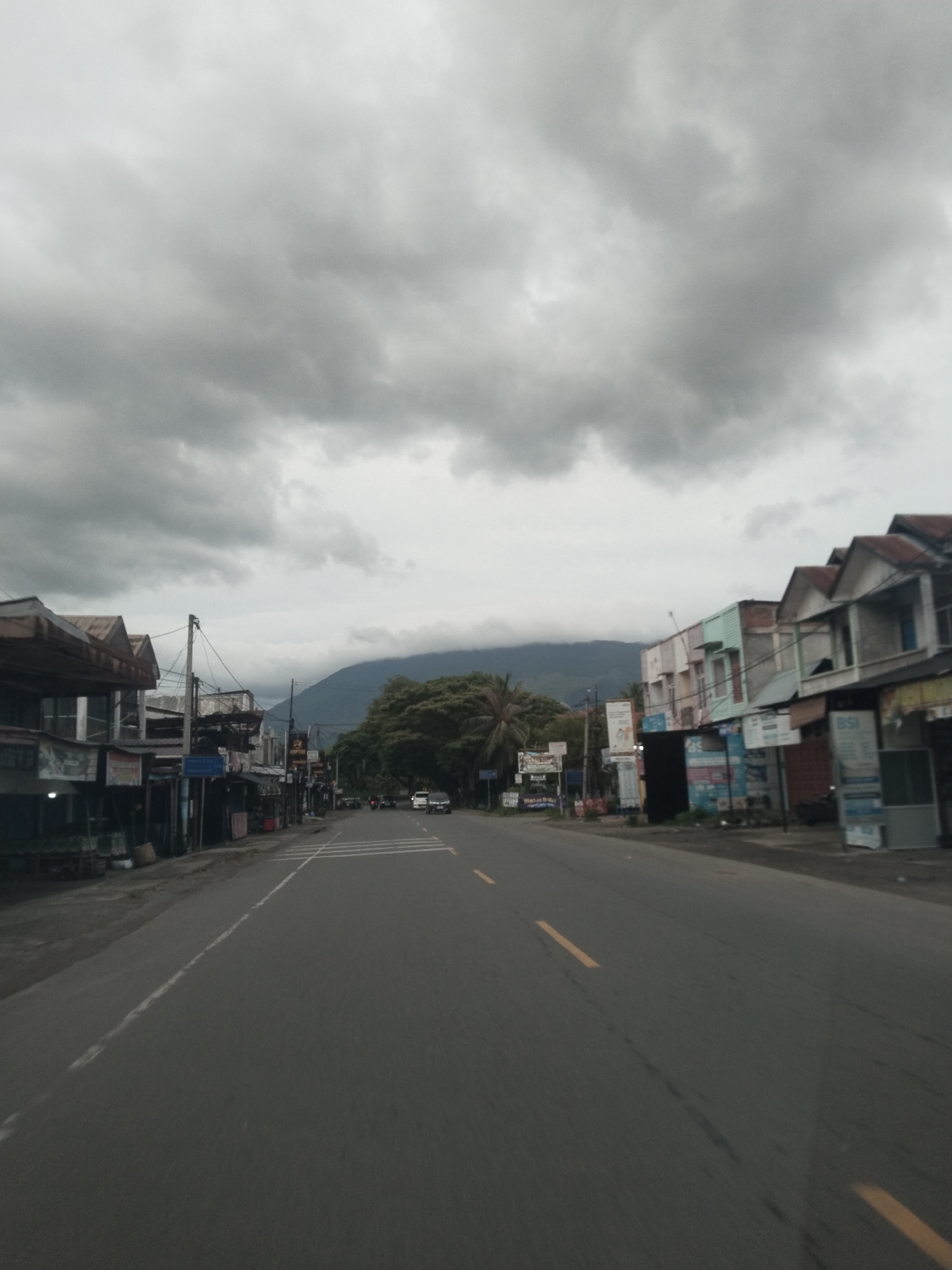 The image depicts a street in Reuhattuha, Indonesia, under an overcast sky. The road has two lanes separated by a yellow dashed line. Buildings line both sides of the street, varying in height and construction style. A mountain is visible in the distance, partially obscured by low-lying clouds. Several cars are present on the road. There are advertisements visible along the street.