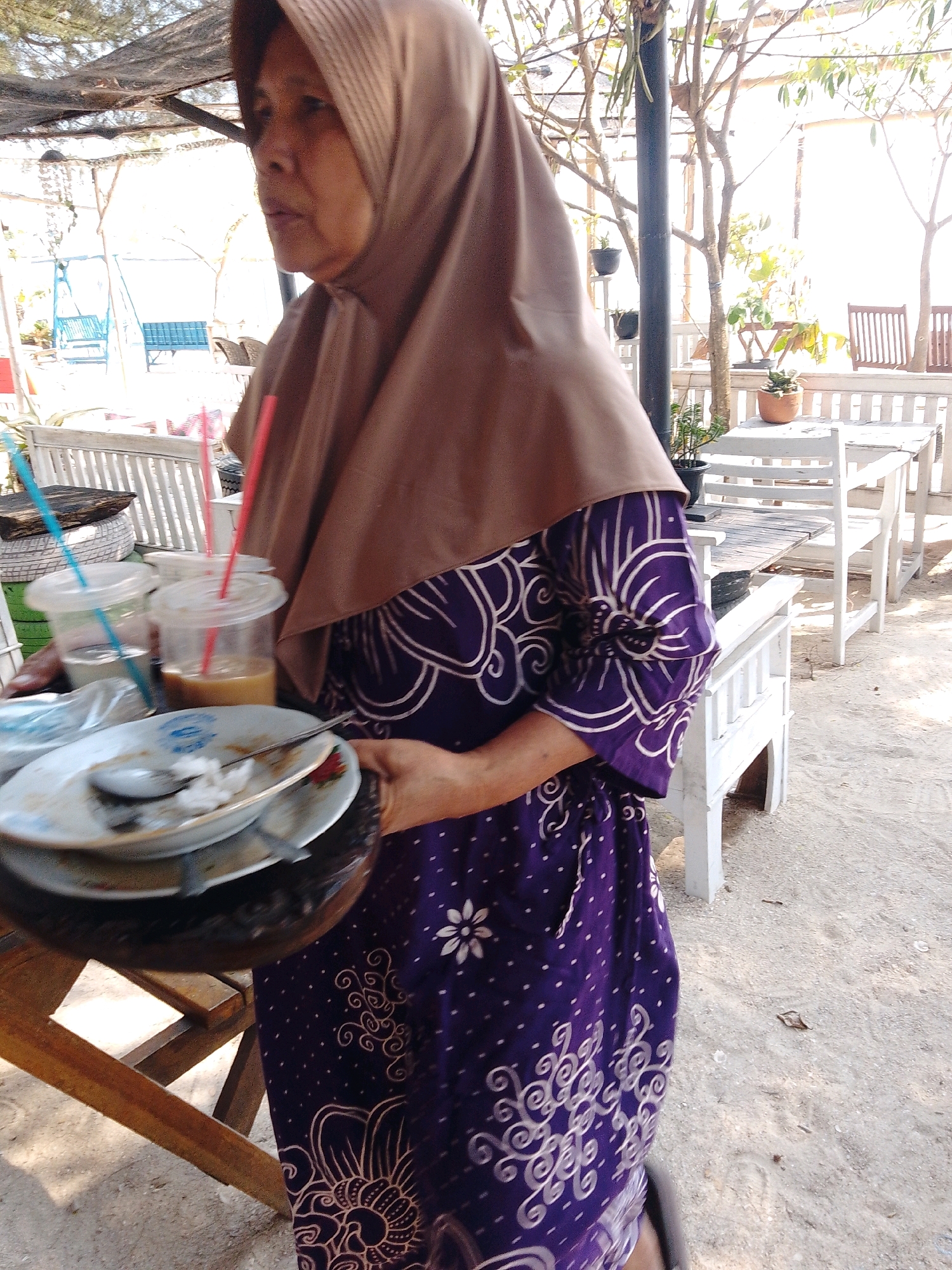 An Indonesian woman wearing a brown hijab and a purple batik dress walks across a sandy area, carrying a tray with two plastic cups and a stack of dirty plates. The cups contain drinks and have straws. The plates are used and have some leftover food and a spoon. The woman is in the foreground, and the background shows an outdoor seating area with white wooden tables and chairs, suggesting a cafe or restaurant. Trees and foliage are visible, along with structures that appear to be part of a shaded seating area. The overall environment suggests a casual, outdoor dining setting in Cikal, Indonesia.