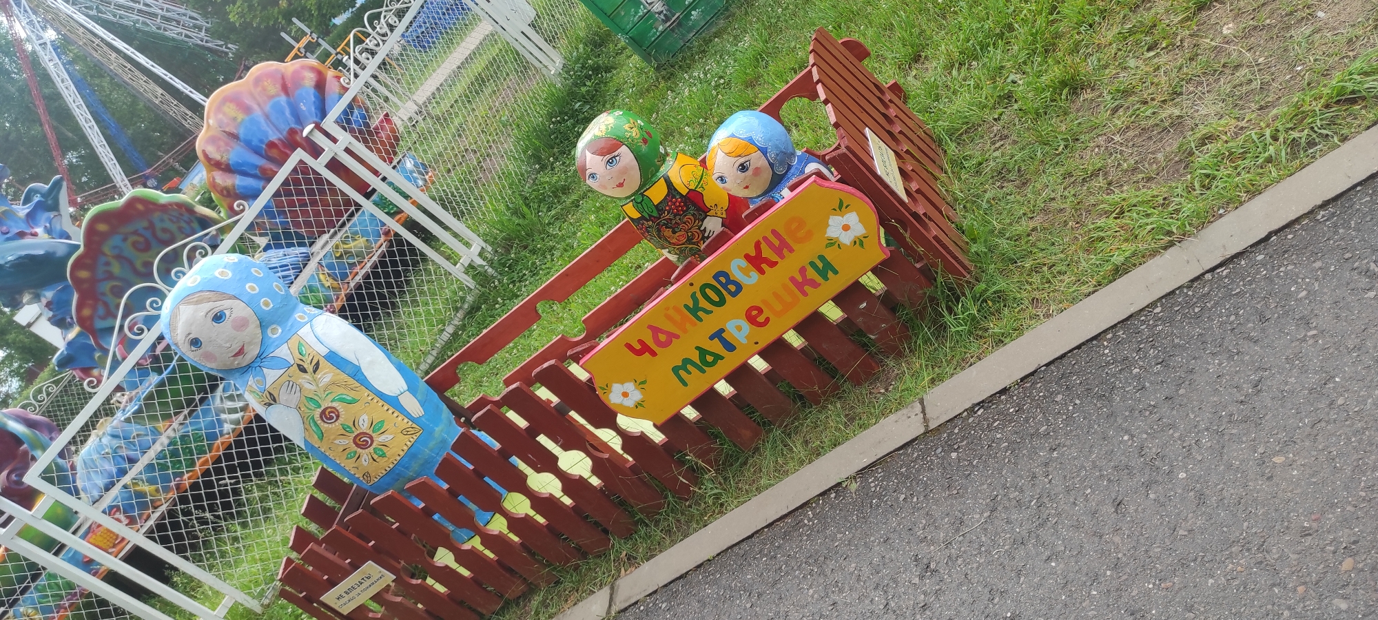 In the city of Chaikovsky, Russia, a photo captures a vibrant scene with a focus on traditional Russian nesting dolls, known as Matryoshka dolls. A large, blue Matryoshka doll with intricate floral patterns stands prominently, positioned behind a rustic red wooden fence.
The fence encloses a grassy area where smaller Matryoshka dolls are displayed. These dolls are depicted with colorful attire and detailed faces, adding to the charm of the exhibit. One doll wears a green headscarf, another a blue one, and they are seated within the fence enclosure.
A sign with the inscription "ЧАЙКОВСКИЕ МАТРЕШКИ" (Chaikovsky Matryoshka dolls) in bright, playful lettering is attached to the fence, further identifying the location and the type of dolls. The sign is adorned with a white flower illustration.
In the background, parts of a colorful amusement park ride are visible, suggesting the location is an entertainment area or park. The ride features a mix of blues, reds, and yellows, with a white metal railing. The overall setting is outdoors, with lush green grass and trees in the distance, indicating a pleasant day. A paved path runs along the edge of the grassy area. A small white sign with some text is affixed to the red fence.