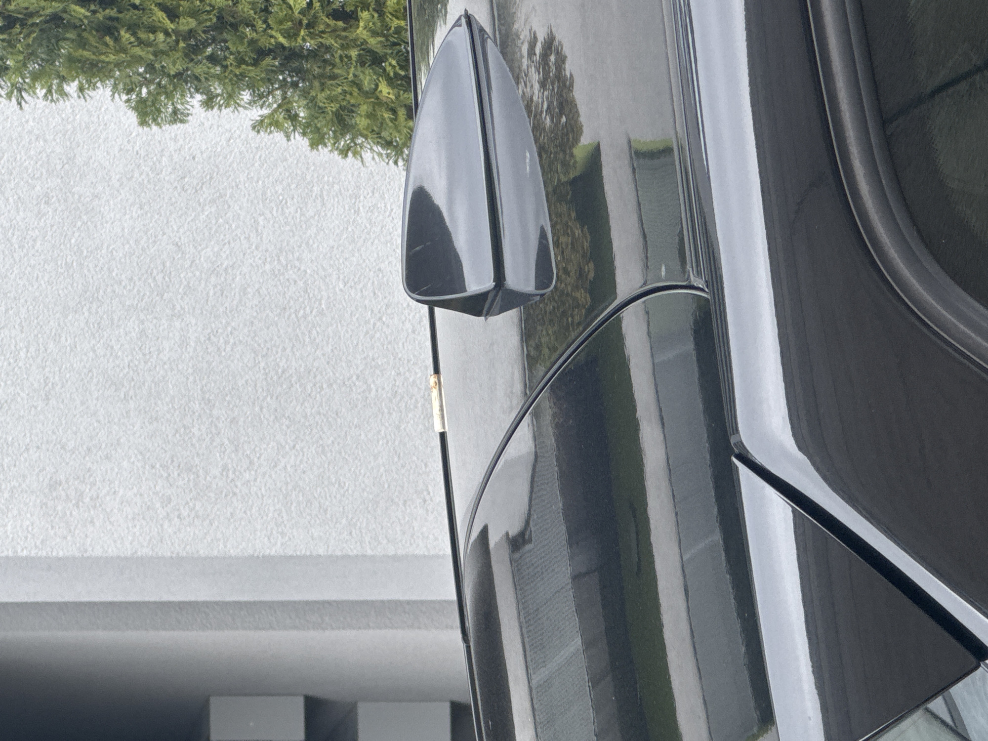 A close-up, angled shot captures a portion of a black vehicle, possibly a car, with reflections visible on its shiny surface. The vehicle features a shark fin antenna. The backdrop consists of a textured white wall and some greenery, which could be trees or bushes. The setting appears to be an urban or suburban area, though specific landmarks are not discernible from the image, so stating the city of Sofia, Bulgaria, is not possible from the context given. The image focuses on the vehicle's details and its immediate surroundings, offering a glimpse into a modern or well-maintained environment. No people are visible in the image.