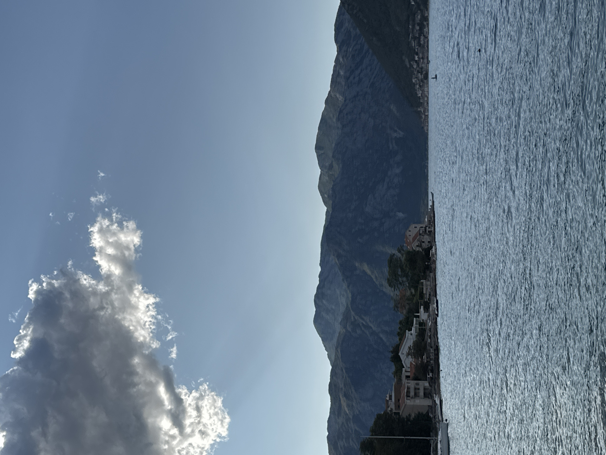 A vertical, slightly high-angle shot captures the waters of the Bay of Kotor in Prčanj, Montenegro, under a partly cloudy sky. In the foreground, the blue water is rippled, reflecting the sky. Along the shore, a cluster of buildings with red-tiled roofs and light-colored facades are visible, interspersed with trees. A white sailboat is moored on the left side of the frame. In the background, a range of dark, rugged mountains slopes down towards the water, their peaks silhouetted against the sky. A few scattered clouds drift across the blue expanse, with sunlight breaking through one larger cloud on the upper left. The overall scene depicts a peaceful coastal view.