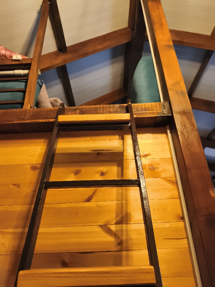 The image depicts an interior scene, primarily focused on a wooden loft structure and its access ladder. A ladder, composed of dark-colored metal side rails and lighter-toned wooden rungs, extends upwards from the lower frame towards a loft platform. The ladder is positioned against a vertical wall constructed from horizontally arranged, light yellowish-brown wooden planks, displaying visible wood grain and knots. Above this wall, a loft area is present, featuring a wooden floor. Within the loft, soft furnishings are visible: a teal-colored cushion or pillow on the right, and a lighter-colored cushion or pillow with a segment of pinkish fabric on the far left. The upper section of the image reveals a sloped ceiling, likely an attic, with light-colored panels (possibly white or light blue) and supported by prominent, dark-stained wooden beams. The overall environment is characterized by natural wood textures and warm illumination, suggesting an indoor setting. No individuals, actions, or specific interactions are discernible. The precise location within Budva, Montenegro, cannot be determined from the visual information provided.
