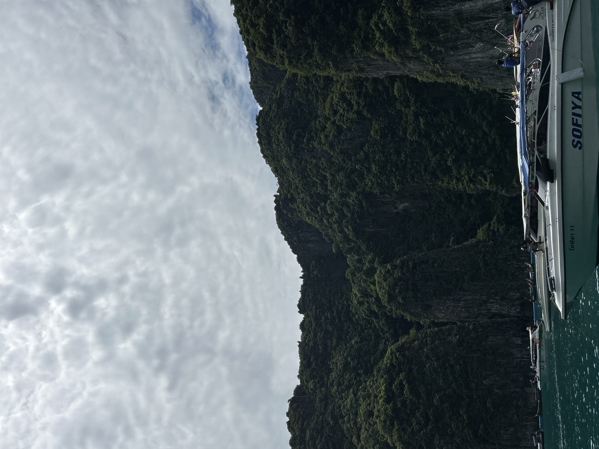 The image depicts a scenic view of towering, lush green cliffs with a body of water in the foreground. Several boats are docked or cruising on the water, with some people visible on board. The sky above is partly cloudy, with a mix of gray and white clouds. The overall impression is one of a tropical coastal landscape, likely a popular tourist destination given the presence of multiple boats. The specific location is identified as Unknown, Thailand.