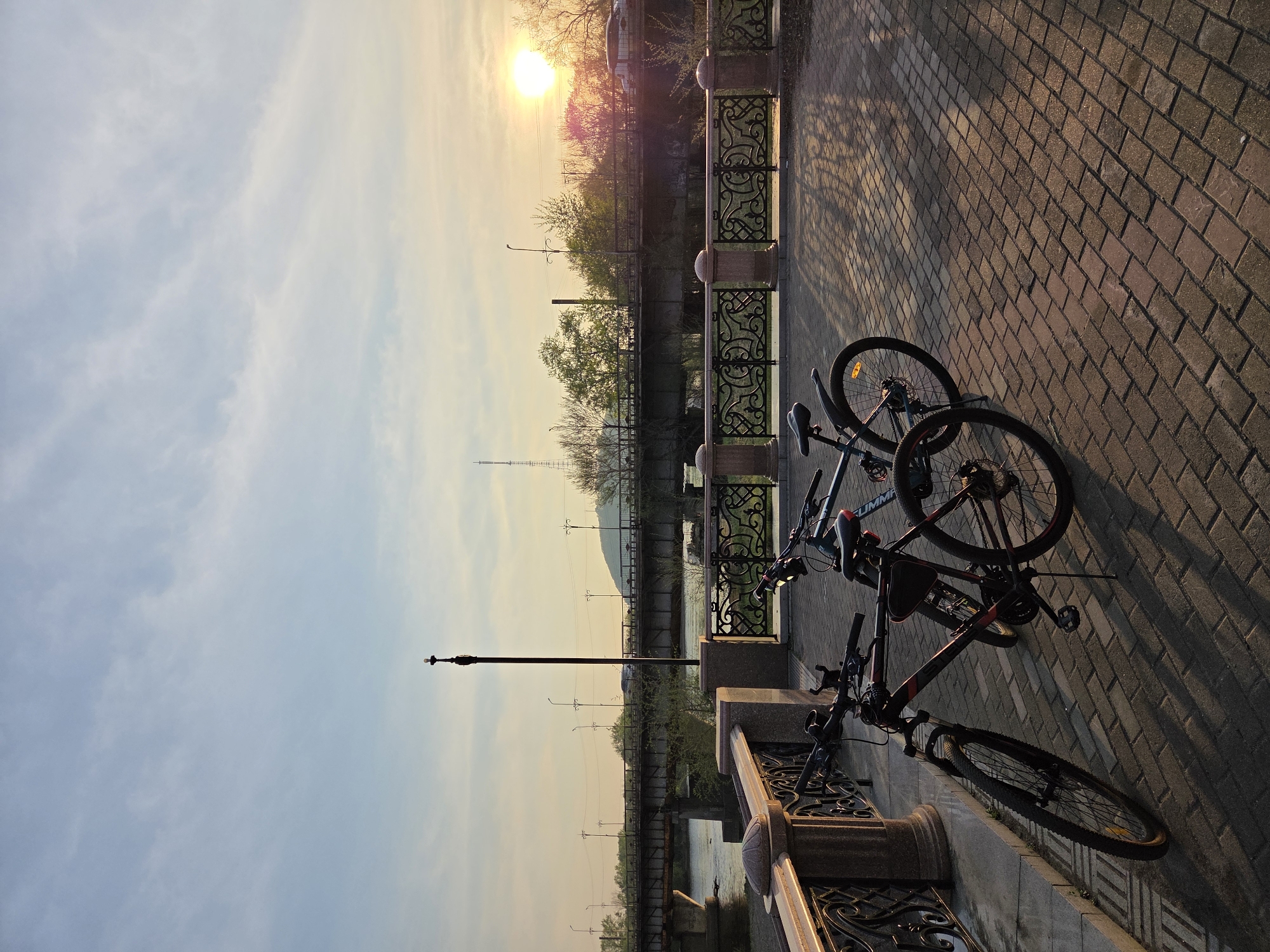 The image captures a serene scene at sunset in what is identified as Birobidzhan, Russia. Two bicycles are parked along an ornate wrought-iron railing that borders a pedestrian walkway. The walkway is paved with brick. In the background, a river can be seen. The sunset casts a warm glow over the scene, illuminating the trees and structures on the opposite side of the river. A tall transmission tower is visible. The sky is a mix of blue and orange hues. The bicycles are the focal point of the image, standing out against the backdrop of the river and sunset.